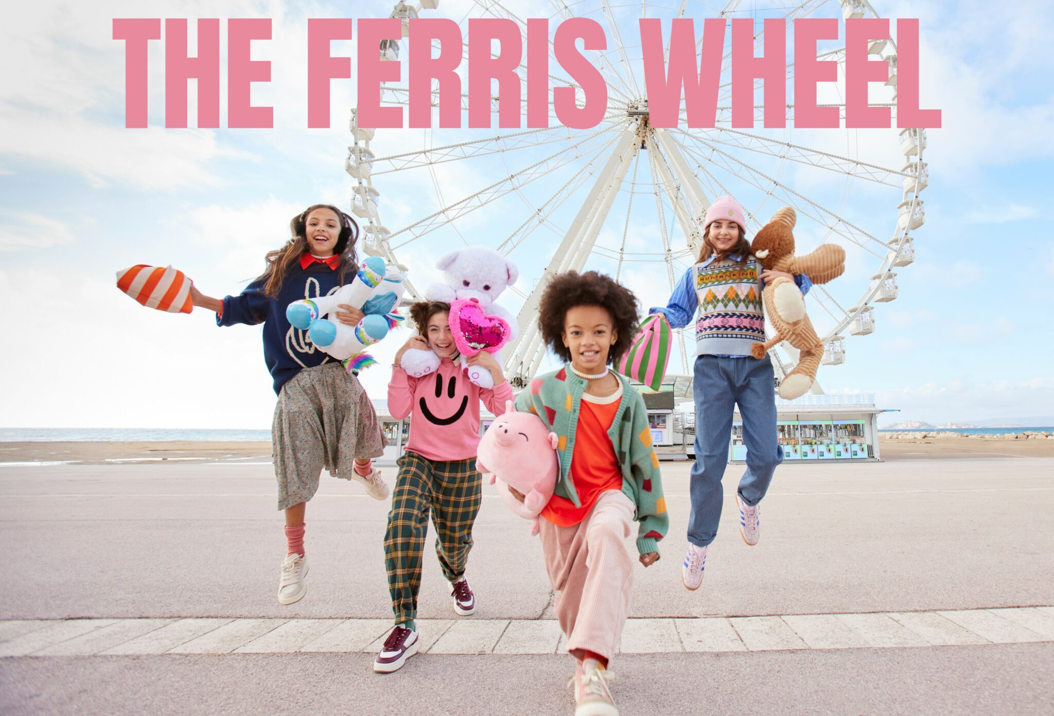 Four children run joyfully toward the camera in front of a large Ferris wheel, each carrying plush toys. They wear bright, playful FW25 outfits as they jump and smile on a seaside promenade. Kids fashion editorial The Ferris Wheel FW25, photographed by Delphine Royer for Scimparello Magazine.