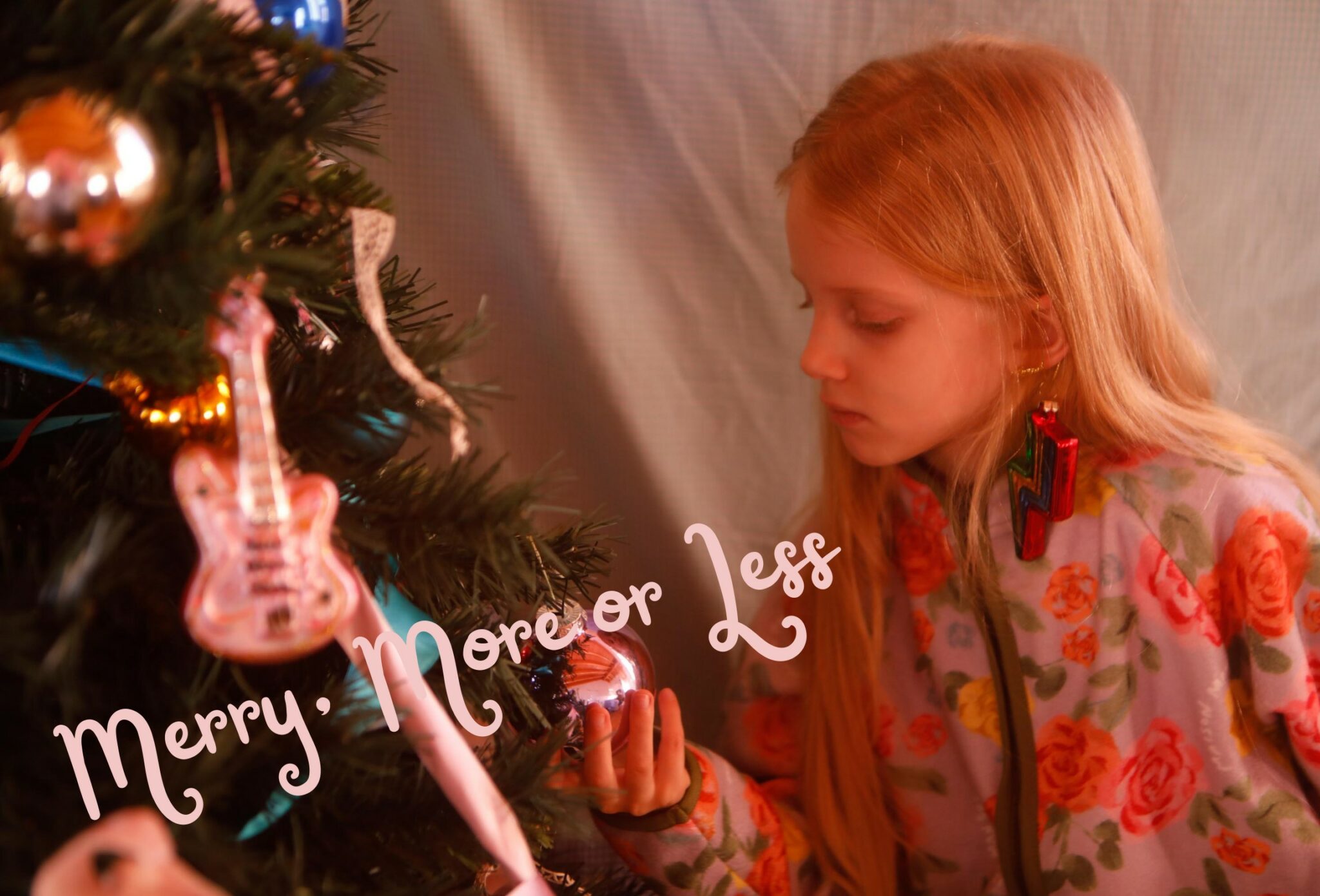 Young girl decorating a Christmas tree with glass ornaments and a guitar-shaped decoration, wearing a floral kids outfit and statement lightning bolt earrings, photographed by Annarella for the kids fashion editorial Merry, more or less.