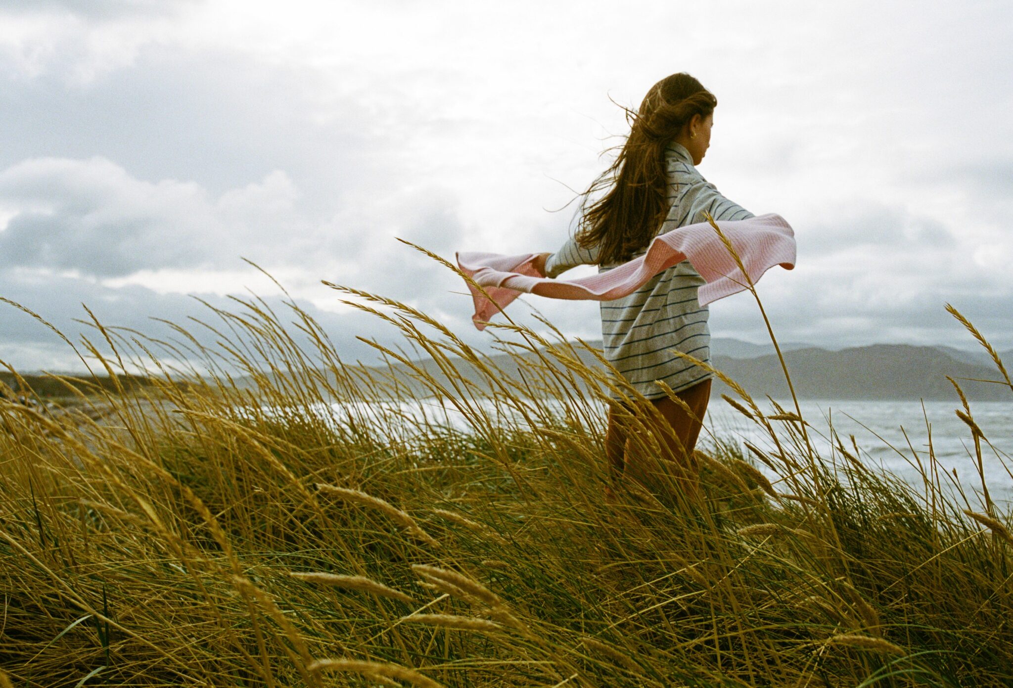Kids fashion editorial Elemental featuring a girl standing in tall coastal grass with arms outstretched, holding a pink scarf, wearing a striped top, with wind-blown hair and the sea in the background, photographed by Hannah Mentz for Scimparello Magazine.
