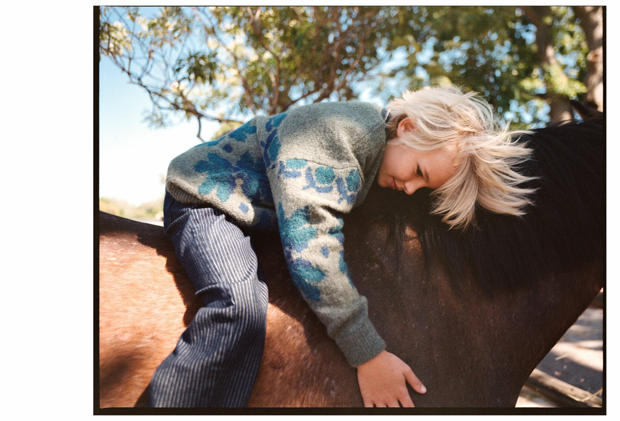 Child lying gently across a pony’s back in a quiet outdoor setting, wearing a patterned knit and striped trousers for the kids fashion editorial Mosi. Photo by Alessandro Iovino, styling by Ulrike Kache, for Scimparello Magazine.