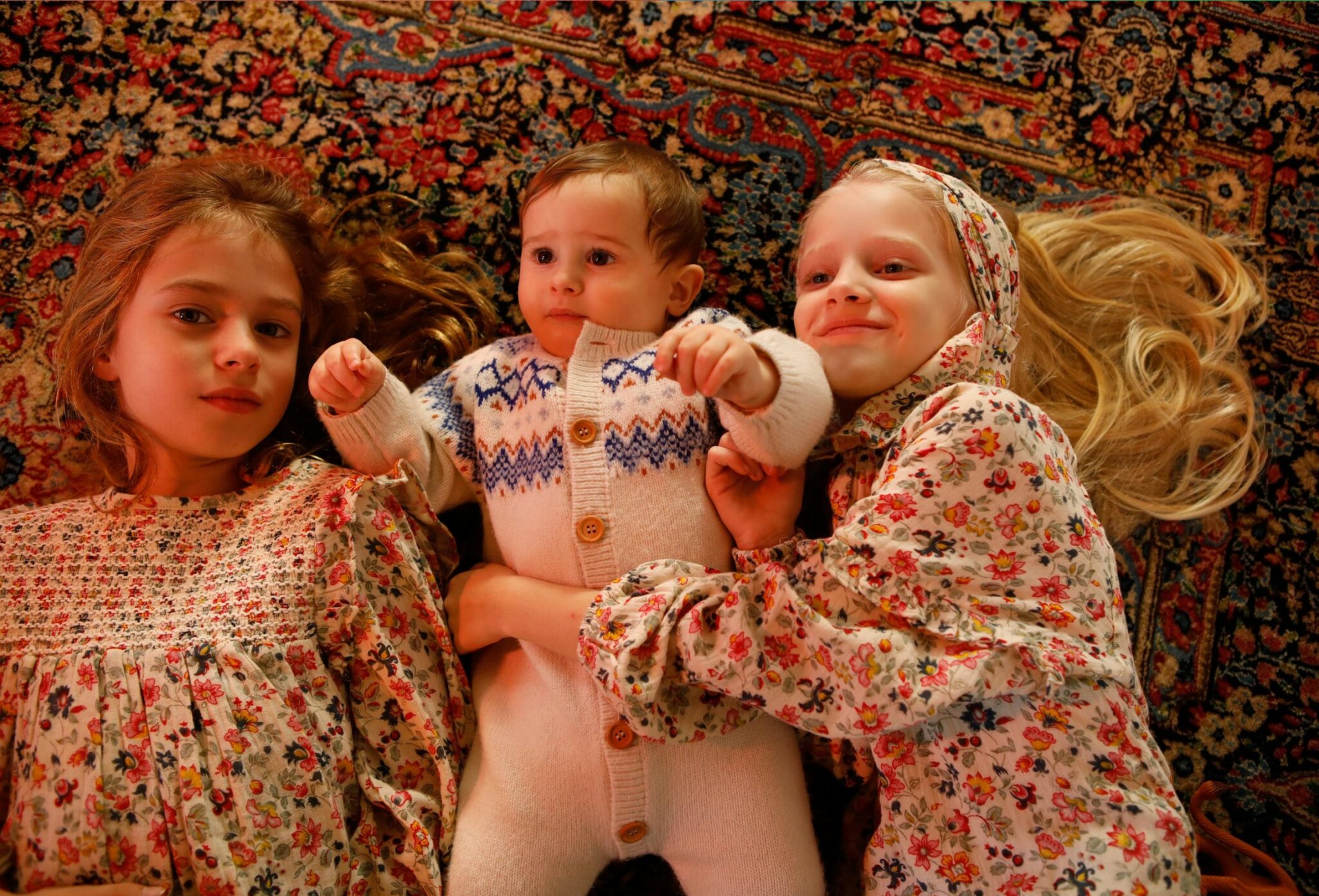 Three children lying close together on a patterned vintage rug, with two girls in floral dresses embracing a baby in knitted romper, captured in a warm, intimate Christmas moment, photographed by Annarella for the kids fashion editorial Merry, more or less for Scimparello Magazine.