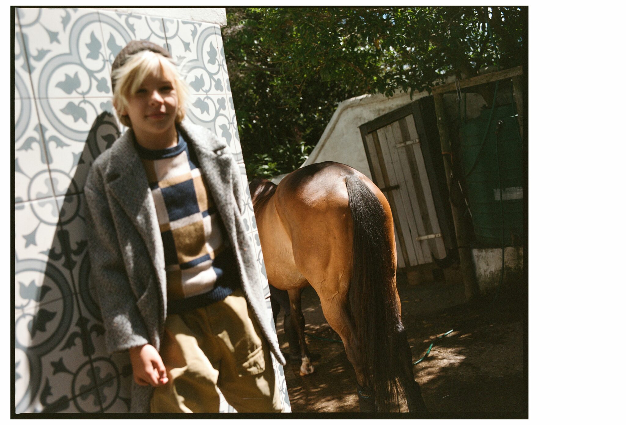 Child standing beside patterned tiles near a stable, with a horse in the background, wearing a layered countryside outfit for the kids fashion editorial Mosi. Photo by Alessandro Iovino, styling by Ulrike Kache, for Scimparello Magazine.