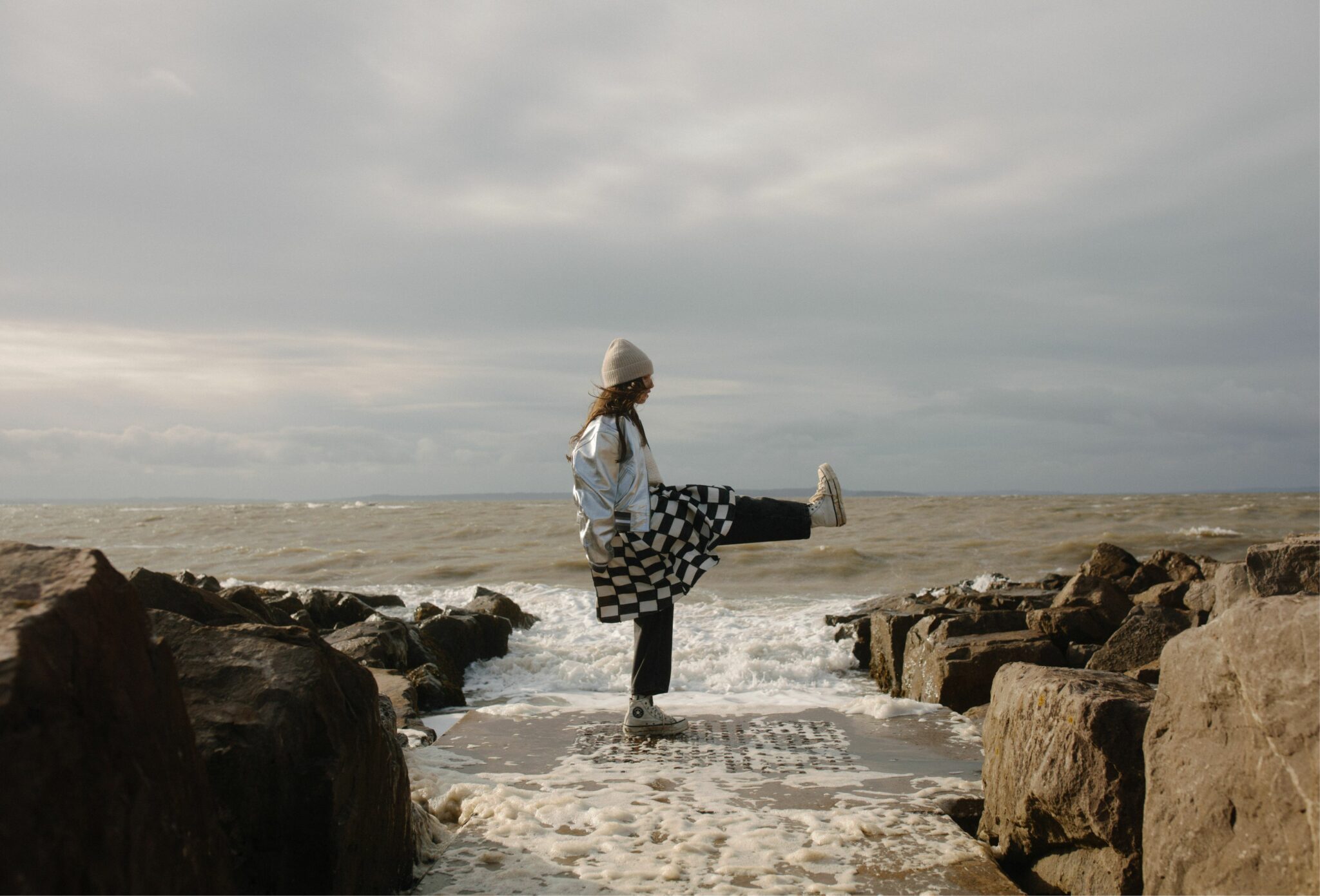 Kids fashion editorial Elemental featuring a girl balancing on a seaside pier with one leg raised, wearing a beanie, metallic jacket, checkered skirt and sneakers, surrounded by rocks and waves, photographed by Hannah Mentz for Scimparello Magazine.