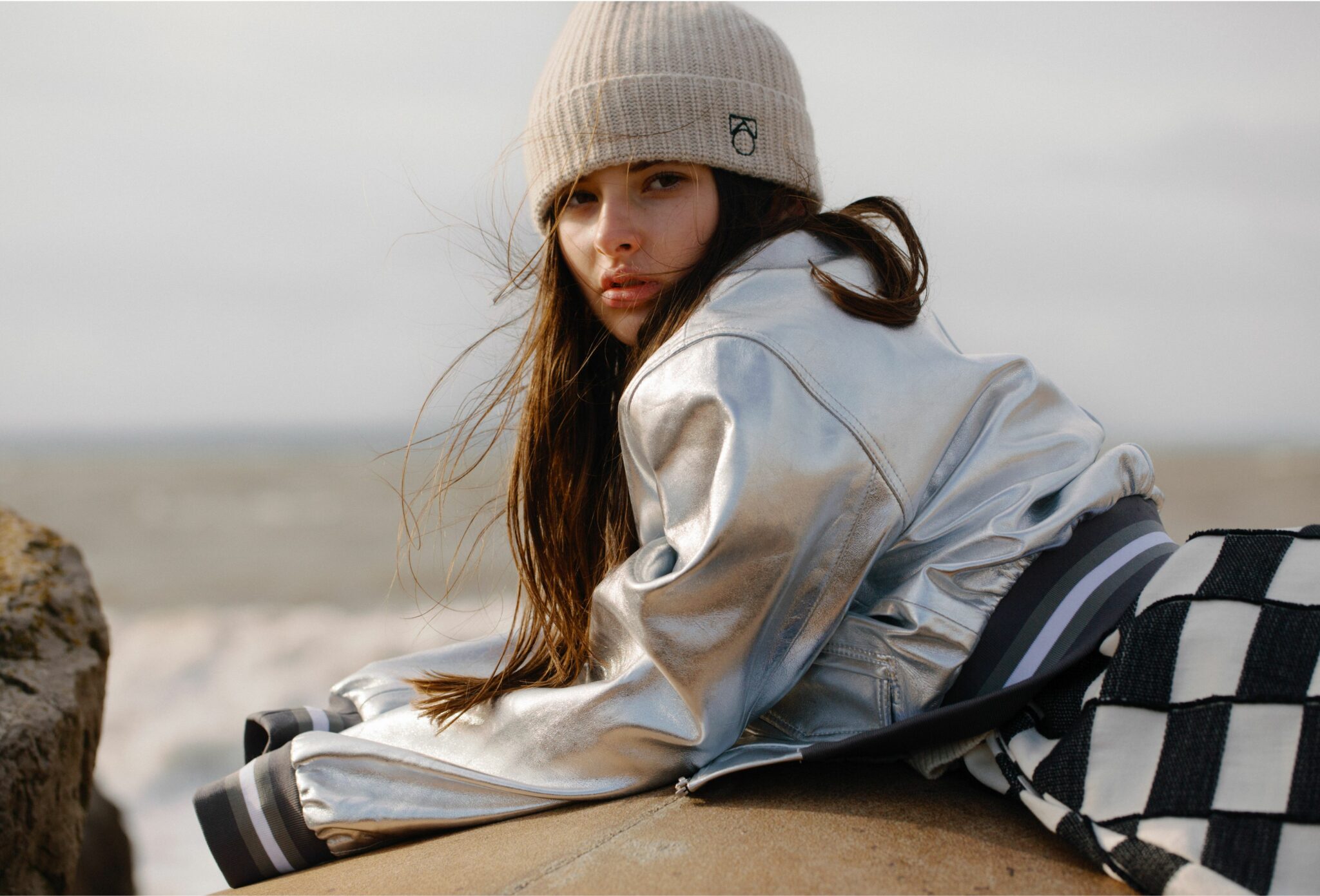 Kids fashion editorial Elemental featuring a close-up of a girl leaning on seaside rocks, wearing a beige beanie, metallic silver jacket and checkered skirt, with wind-blown hair and the sea in the background, photographed by Hannah Mentz for Scimparello Magazine.