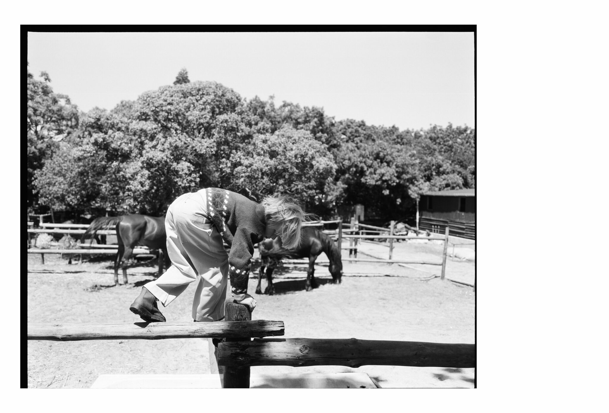 Black and white image of a child balancing on a wooden fence in a paddock, with horses grazing in the background, captured in a quiet rural moment for the kids fashion editorial Mosi. Photo by Alessandro Iovino, styling by Ulrike Kache, for Scimparello Magazine.