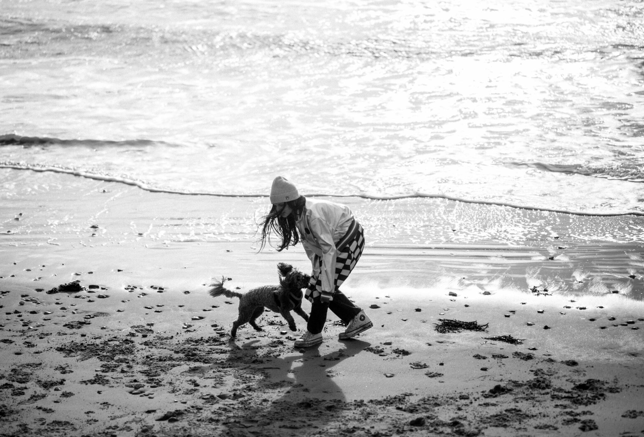 Black and white kids fashion editorial Elemental featuring a girl playing with a small dog on a sandy beach by the shoreline, wearing a beanie, jacket and checkered skirt, with waves in the background, photographed by Hannah Mentz for Scimparello Magazine.