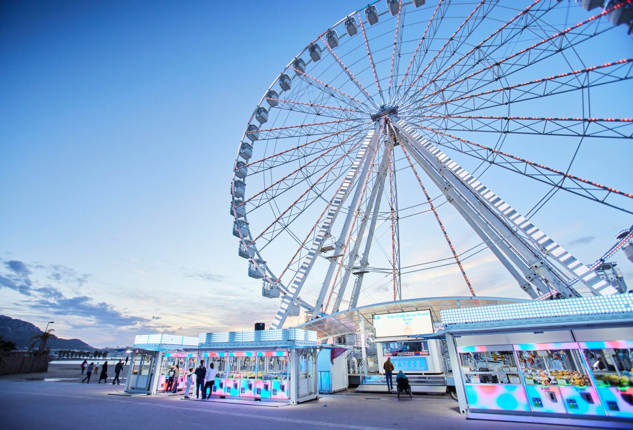 Wide-angle photo of a large Ferris wheel at dusk, illuminated with colourful lights against a blue and pink sky. Small groups of people walk near the ticket booths and game stands below, creating a lively seaside atmosphere.
The Ferris Wheel, kids fashion editorial FW25 photographed by Delphine Royer for Scimparello Magazine.