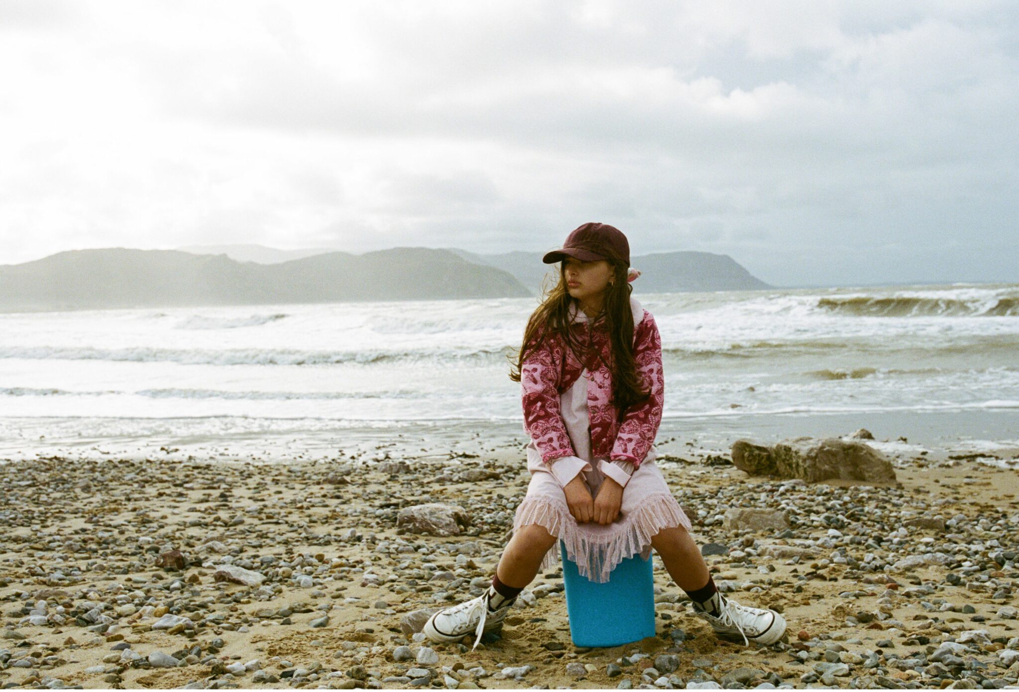 Kids fashion editorial Elemental featuring a girl sitting on a blue block on a pebble beach, wearing a pink patterned jacket, fringed dress, baseball cap and sneakers, with waves and distant hills in the background, photographed by Hannah Mentz for Scimparello Magazine.