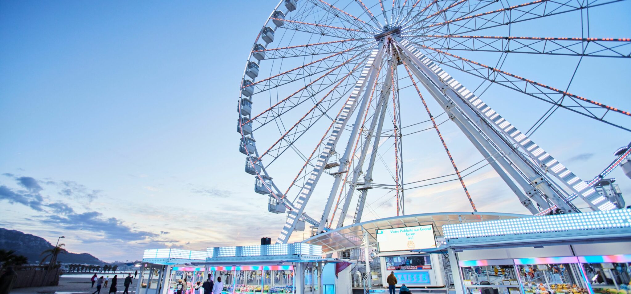 Wide-angle photo of a large Ferris wheel at dusk, illuminated with colourful lights against a blue and pink sky. Small groups of people walk near the ticket booths and game stands below, creating a lively seaside atmosphere. The Ferris Wheel, kids fashion editorial FW25 photographed by Delphine Royer for Scimparello Magazine.