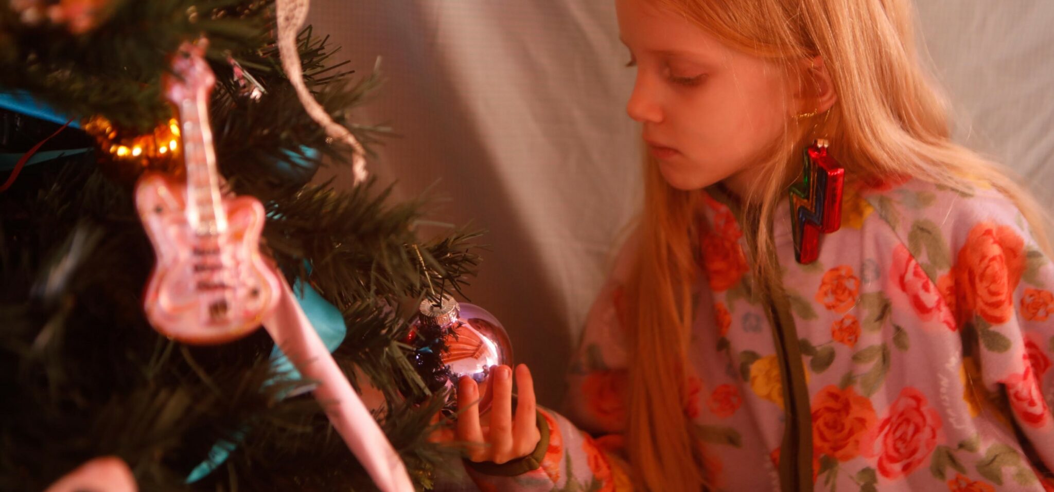 Young girl decorating a Christmas tree with glass ornaments and a guitar-shaped decoration, wearing a floral kids outfit and statement lightning bolt earrings, photographed by Annarella for the kids fashion editorial Merry, more or less.