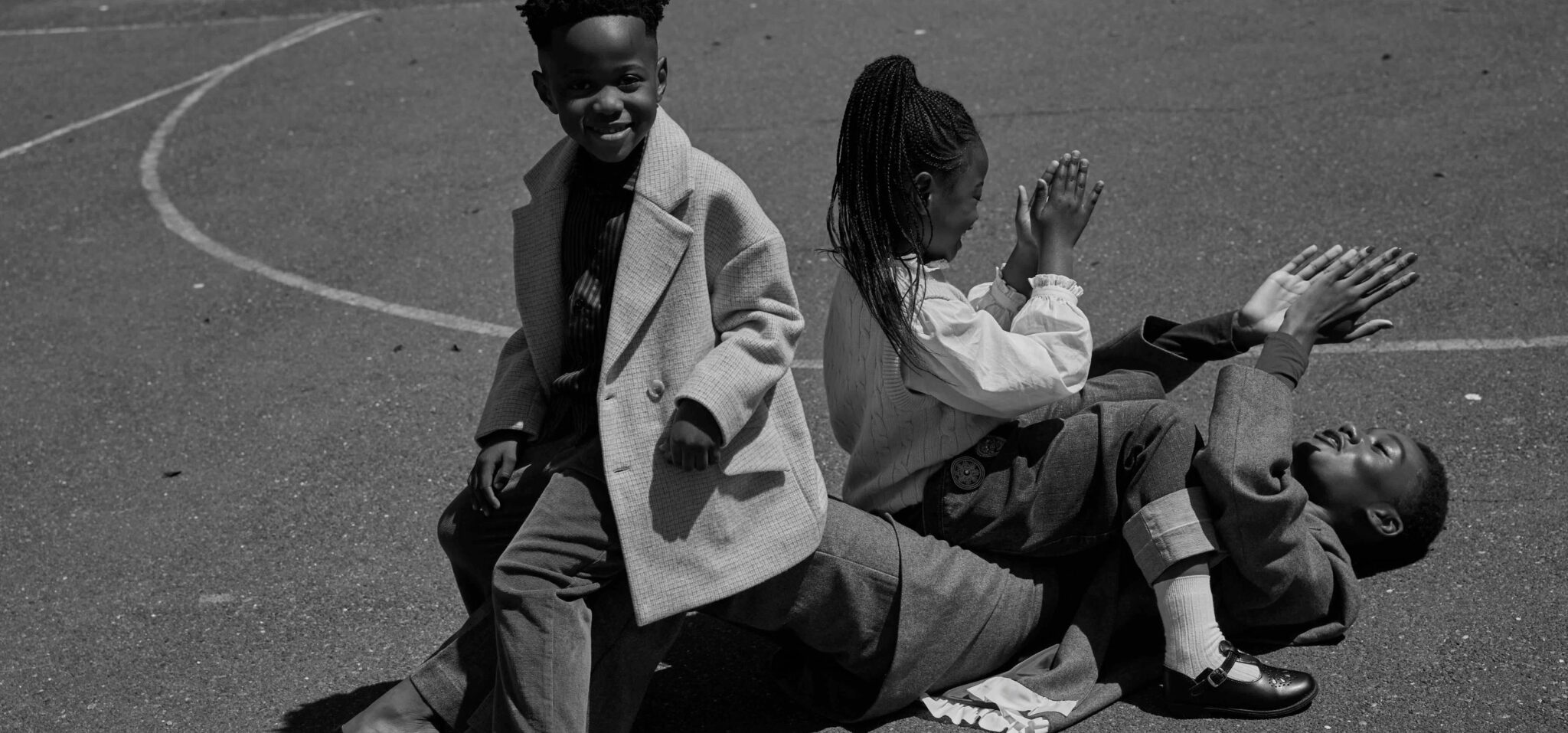 Black-and-white photo of two children sitting on a playground basketball court, leaning back against a woman who is lying on the floor behind them. The scene feels relaxed and playful.