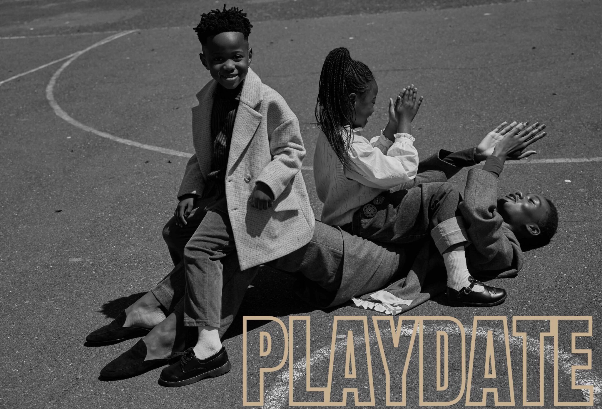 Black-and-white photo of two children sitting on a playground basketball court, leaning back against a woman who is lying on the floor behind them. The scene feels relaxed and playful.