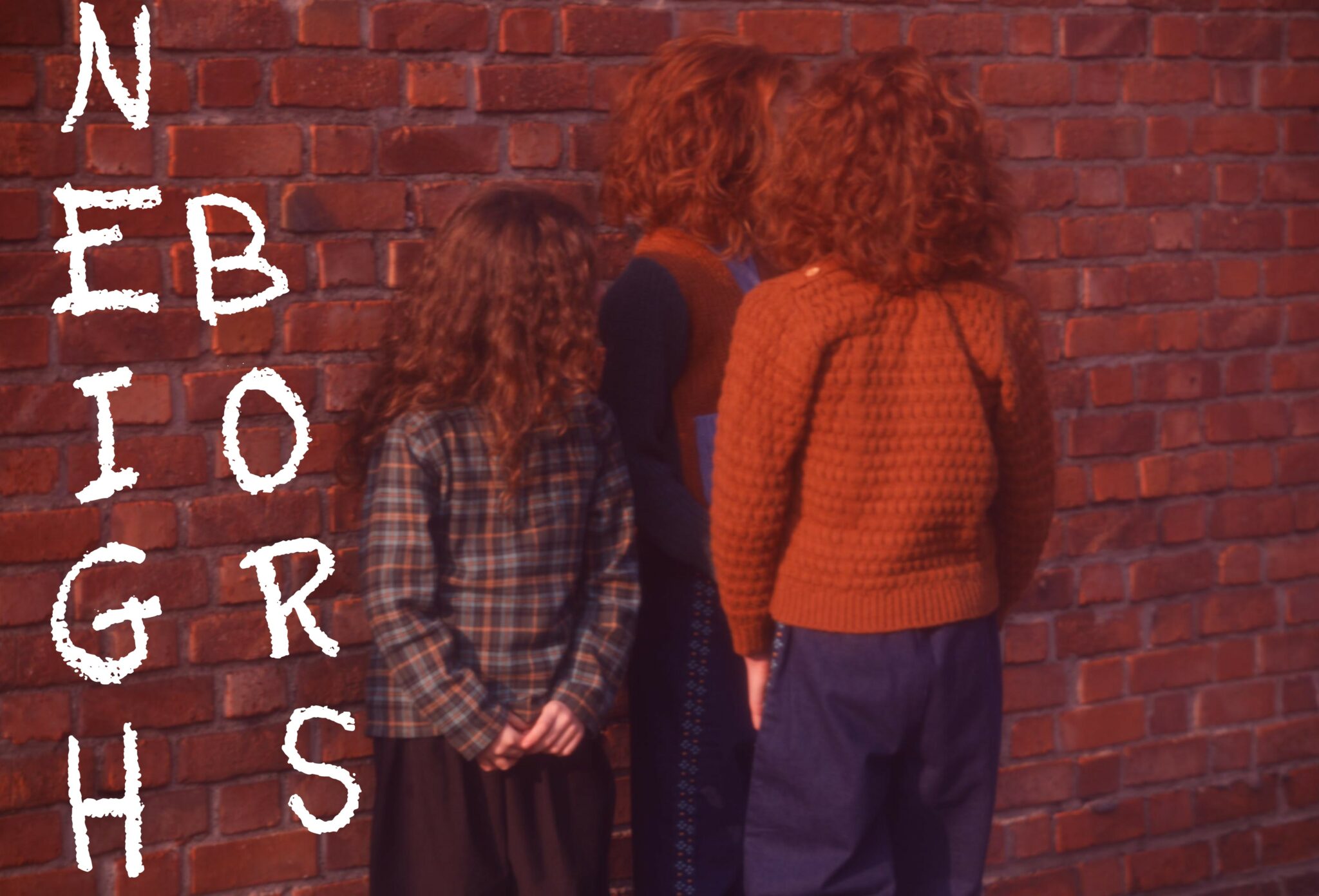 Kids fashion editorial NEIGHBORS for Scimparello Magazine featuring three children seen from behind, standing close to a red brick wall, wearing textured knitwear and layered winter outfits in warm earthy tones. A quiet, nostalgic moment captured in a city setting. Photo by Annarella Caruso.