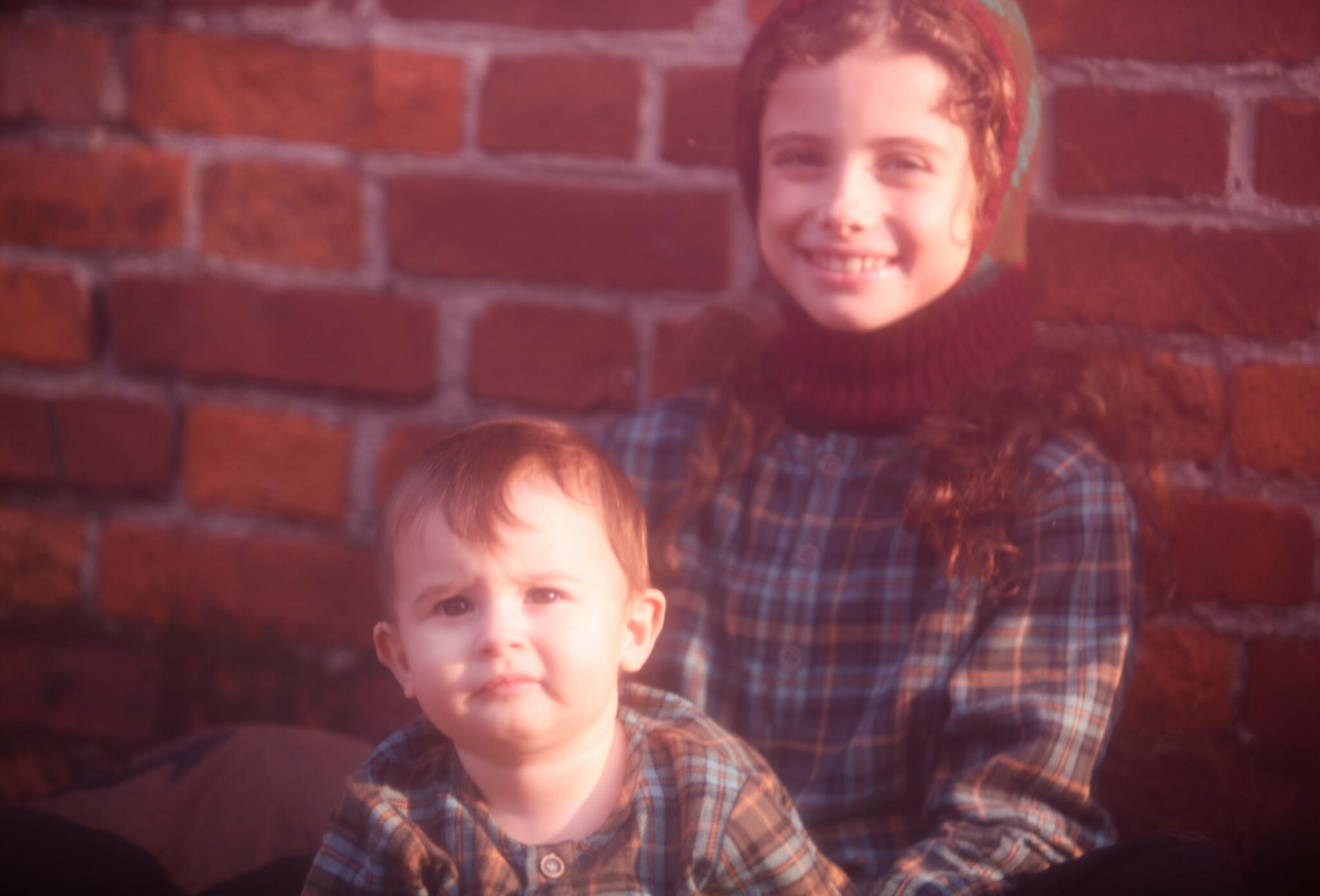 Kids fashion editorial NEIGHBORS for Scimparello Magazine featuring two children sitting in front of a red brick wall, wearing matching plaid winter outfits in warm earthy tones. A soft, intimate portrait capturing sibling closeness and everyday childhood moments. Photo by Annarella Caruso.