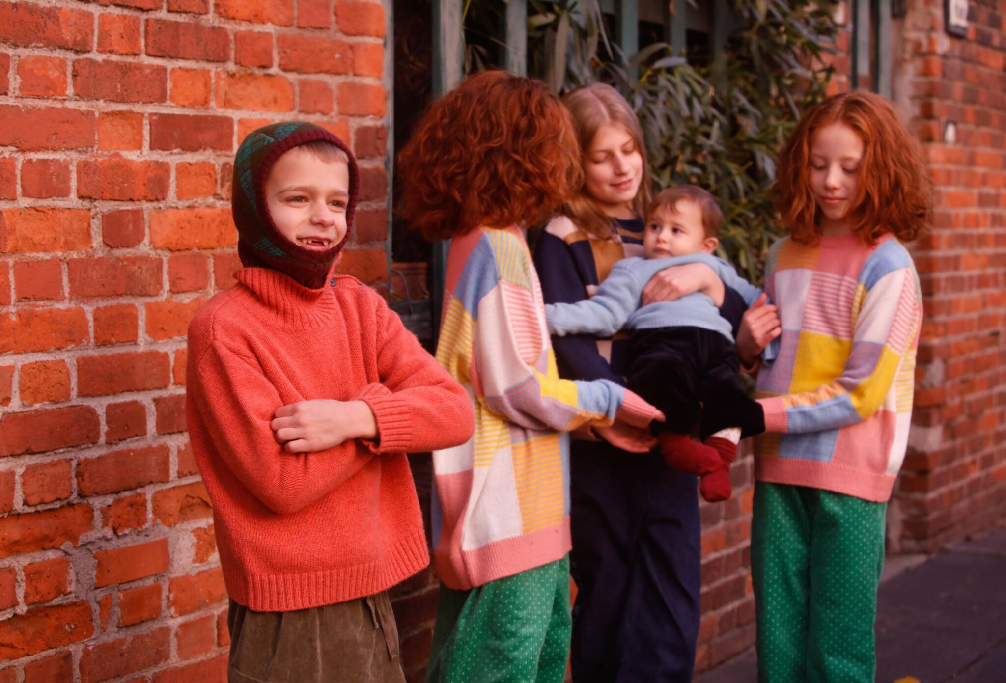 Kids fashion editorial NEIGHBORS for Scimparello Magazine featuring a group of children and a baby standing together beside a red brick wall, wearing colorful knitted sweaters and winter layers. A spontaneous moment capturing closeness, play, and everyday childhood life. Photo by Annarella Caruso.