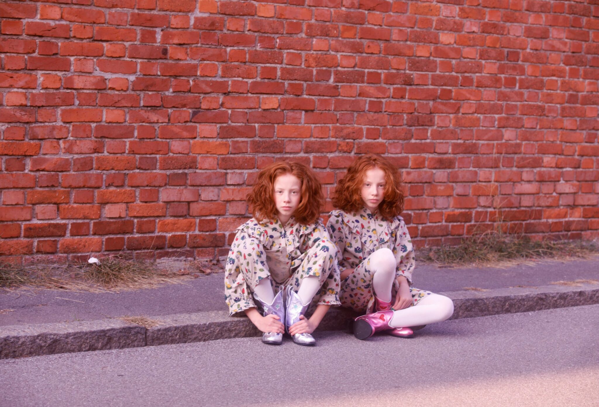 Kids fashion editorial NEIGHBORS for Scimparello Magazine featuring two red-haired girls sitting on a curb in front of a red brick wall, wearing matching floral dresses, white tights, and metallic cowboy boots. A quiet, reflective moment capturing individuality and everyday childhood style. Photo by Annarella Caruso.