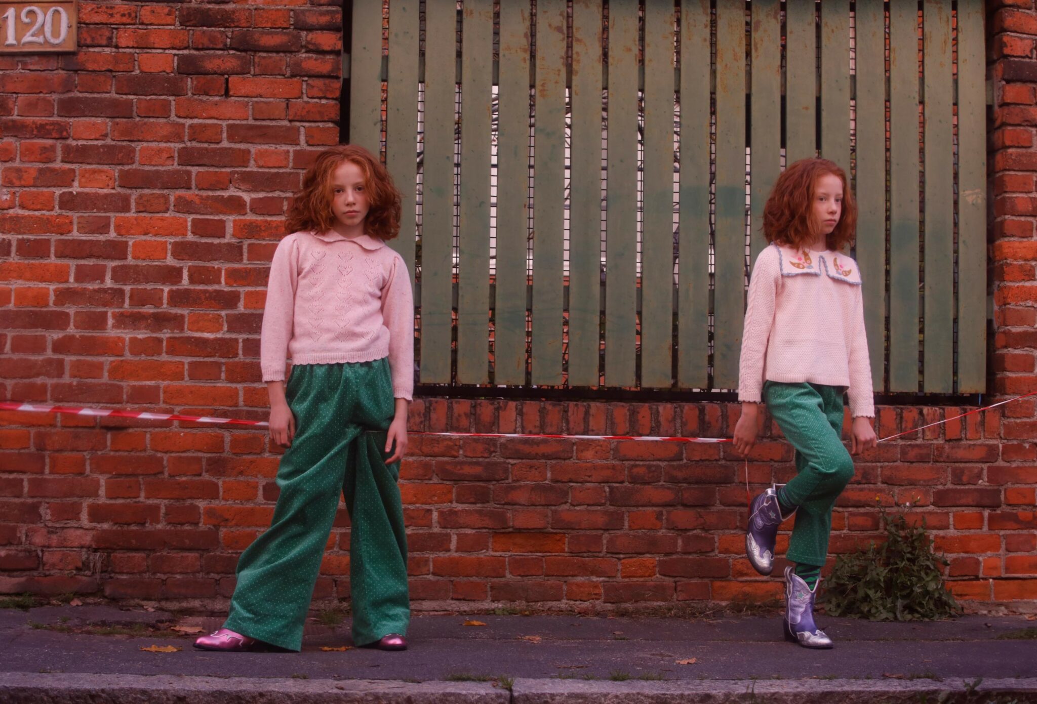 Kids fashion editorial NEIGHBORS for Scimparello Magazine featuring two red-haired twins standing apart along a brick wall and green wooden fence, wearing soft pink knit sweaters, green trousers, and metallic boots. A quiet urban scene exploring individuality, distance, and everyday childhood style. Photo by Annarella Caruso.