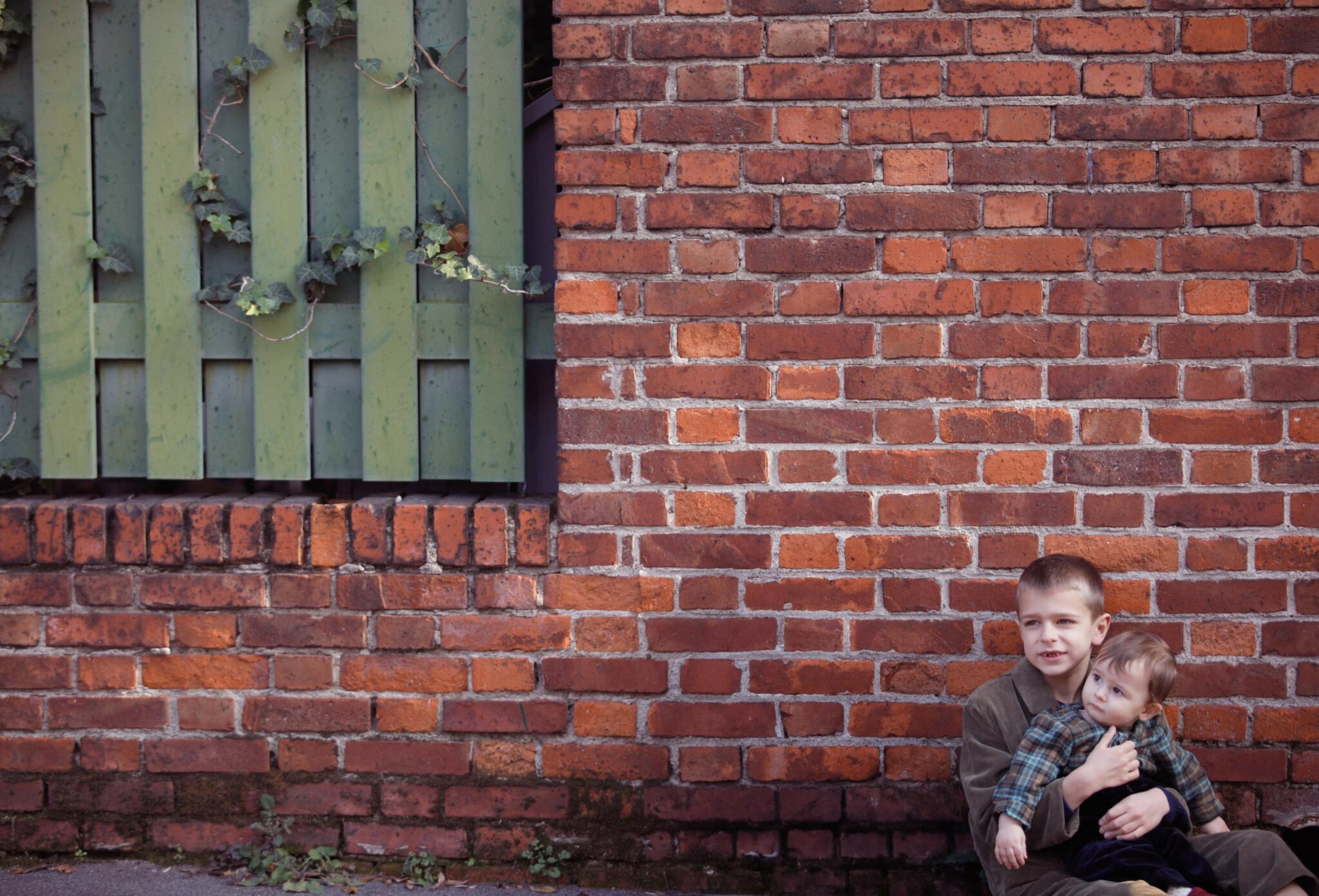 Kids fashion editorial NEIGHBORS for Scimparello Magazine featuring two young siblings sitting together against a red brick wall, one holding the other in a quiet, protective gesture. A calm urban moment capturing closeness, care, and everyday childhood life. Photo by Annarella Caruso.