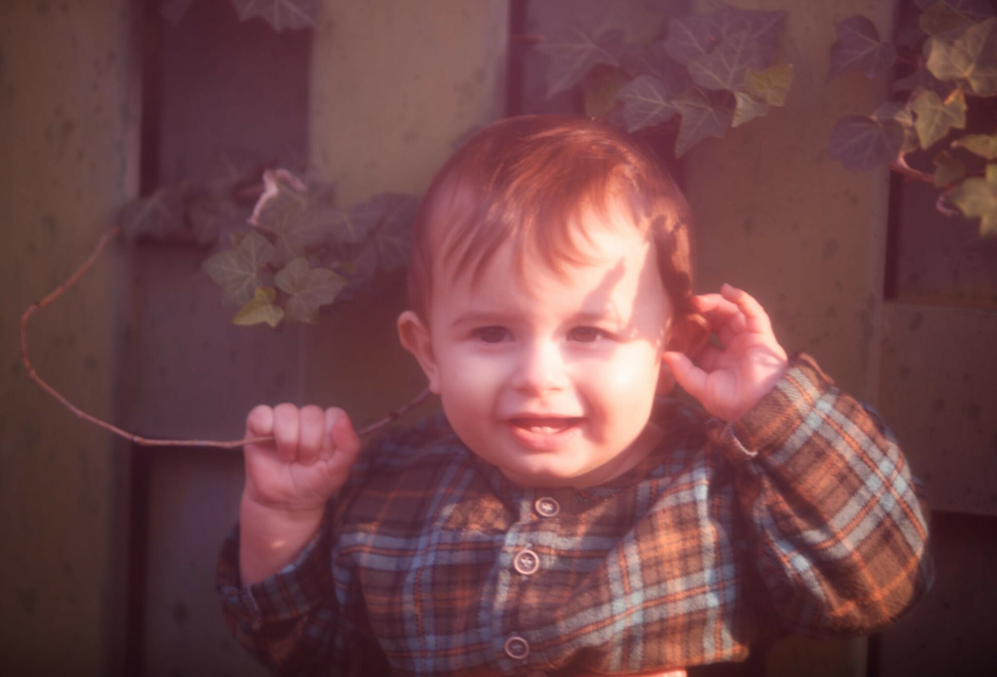 Kids fashion editorial NEIGHBORS for Scimparello Magazine featuring a baby wearing a soft plaid outfit, holding a small branch and smiling in warm afternoon light. A gentle portrait capturing curiosity, innocence, and everyday childhood poetry. Photo by Annarella Caruso.
