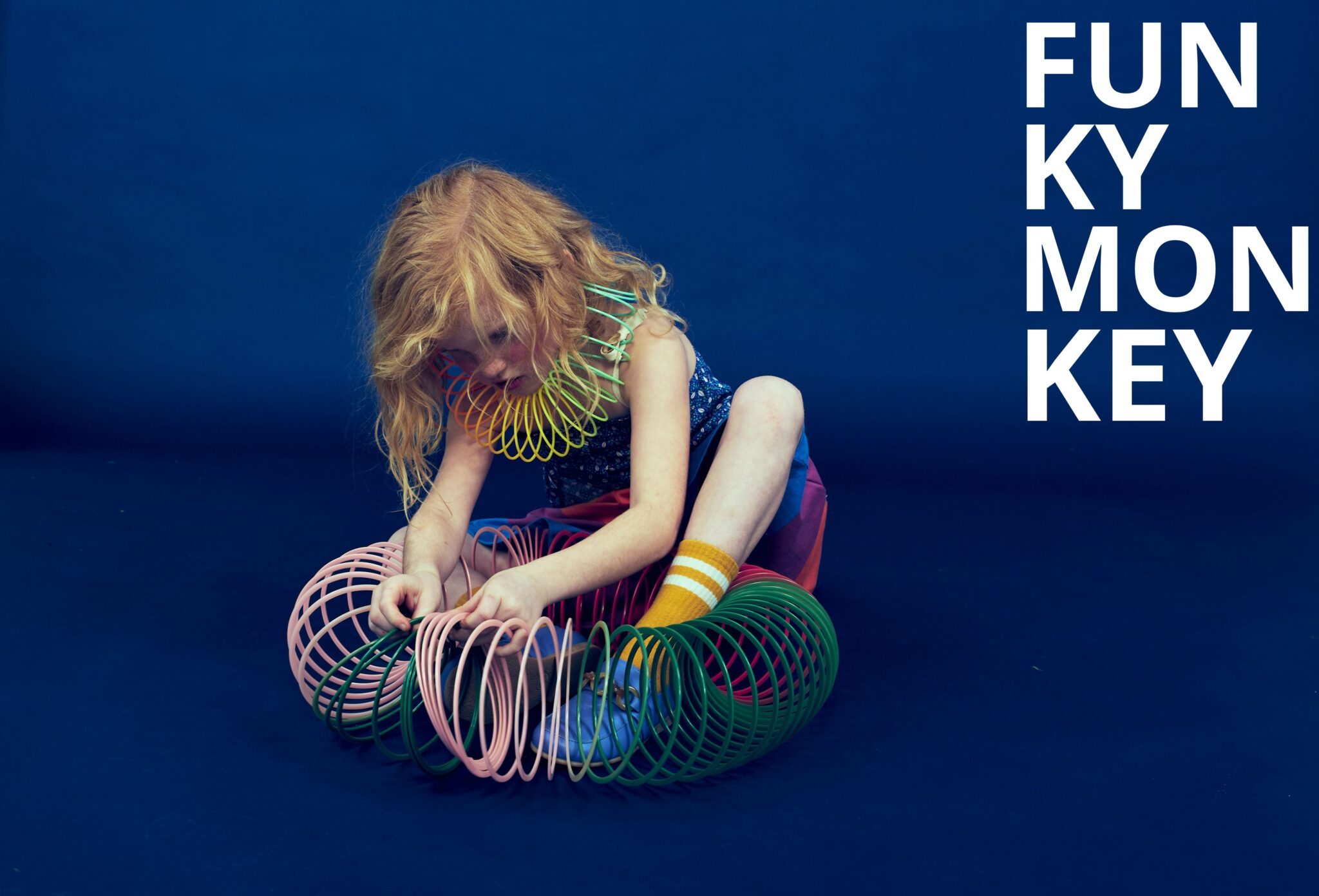 Kids fashion editorial Funky Monkey for Scimparello Magazine featuring a red-haired child sitting on a deep blue studio floor, playing with colorful plastic slinky toys wrapped around arms and legs, wearing a patterned sleeveless top, multicolor skirt and yellow striped socks. Photo by Valerie Mathilde.