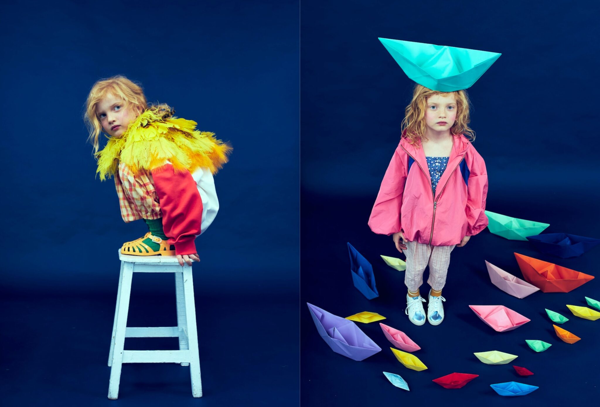 Kids fashion editorial Funky Monkey for Scimparello Magazine featuring a red-haired child in a deep blue studio: on the left, crouching on a white stool wearing a yellow feather collar, checked top, red sweatshirt and green socks with yellow sandals; on the right, standing in a pink oversized jacket and light trousers surrounded by colorful paper boats with a large turquoise paper boat balanced on the head. Photo by Valerie Mathilde.