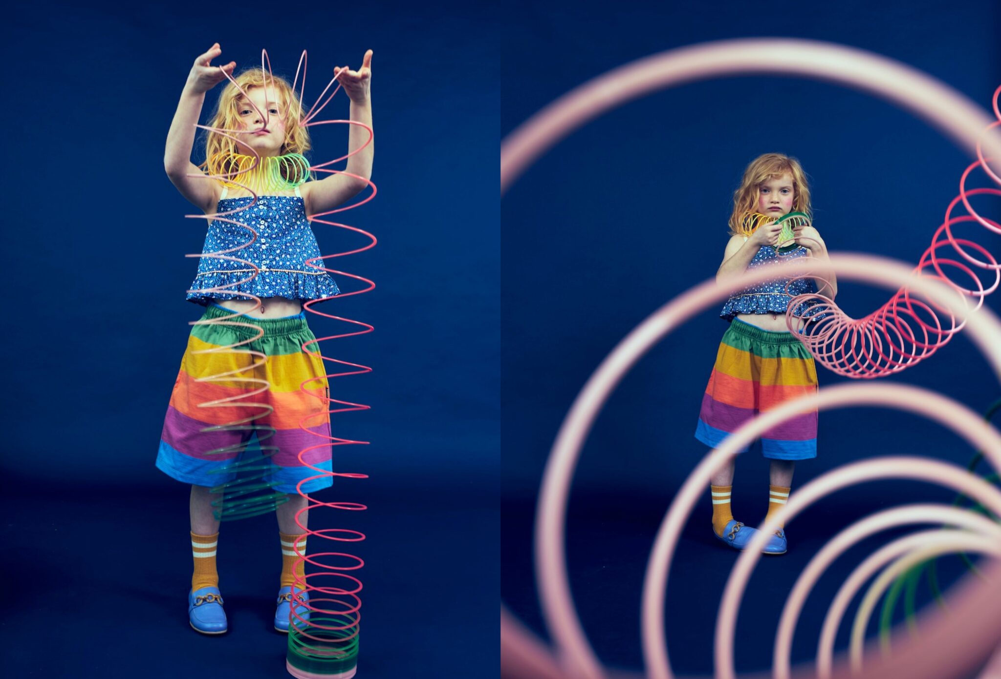 Kids fashion editorial Funky Monkey for Scimparello Magazine featuring a red-haired child in a deep blue studio wearing a blue floral sleeveless top and a rainbow-striped shorts with yellow socks and blue loafers, playing with colorful plastic slinky toys that spiral around the body and frame the foreground. Photo by Valerie Mathilde.