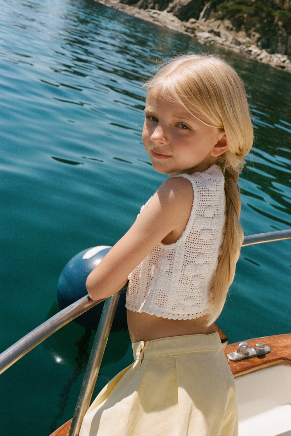 Bebe Organic SS26 blonde girl in crochet top and soft yellow skirt leaning on a boat railing over turquoise water, Mediterranean summer setting, featured on Scimparello Magazine.