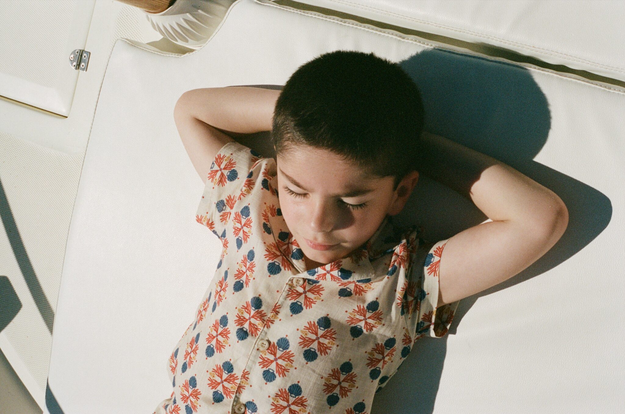 Bebe Organic SS26 boy in coral and blue shell print summer set lying on boat cushion under warm sunlight, relaxed nautical vibe, featured on Scimparello Magazine.
