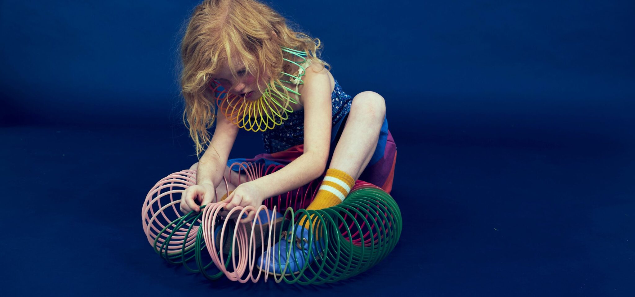 Kids fashion editorial Funky Monkey for Scimparello Magazine featuring a red-haired child sitting on a deep blue studio floor, playing with colorful plastic slinky toys wrapped around arms and legs, wearing a patterned sleeveless top, multicolor skirt and yellow striped socks. Photo by Valerie Mathilde.