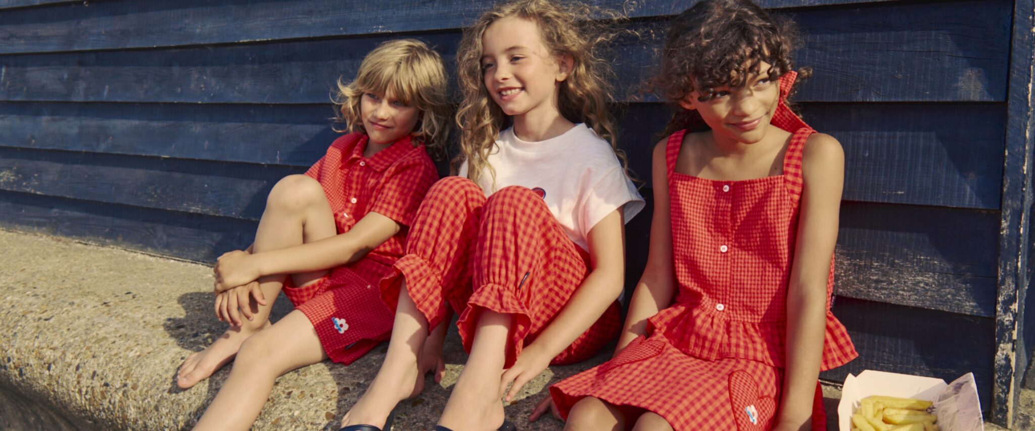 Three children wearing red gingham LOUD outfits sitting against dark blue wooden wall in sunny outdoor setting, LOUD SS26 kids fashion collection featured in Scimparello Magazine.