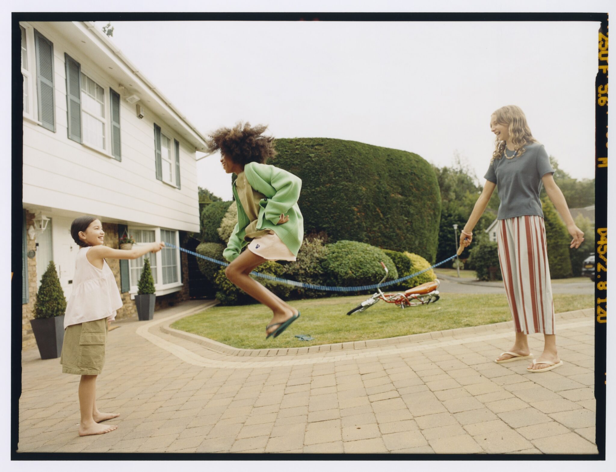 Children playing jump rope in a quiet suburban driveway, girl mid-air wearing green cardigan and pastel outfit, MAIN STORY SS26 kids fashion editorial featured in Scimparello Magazine.