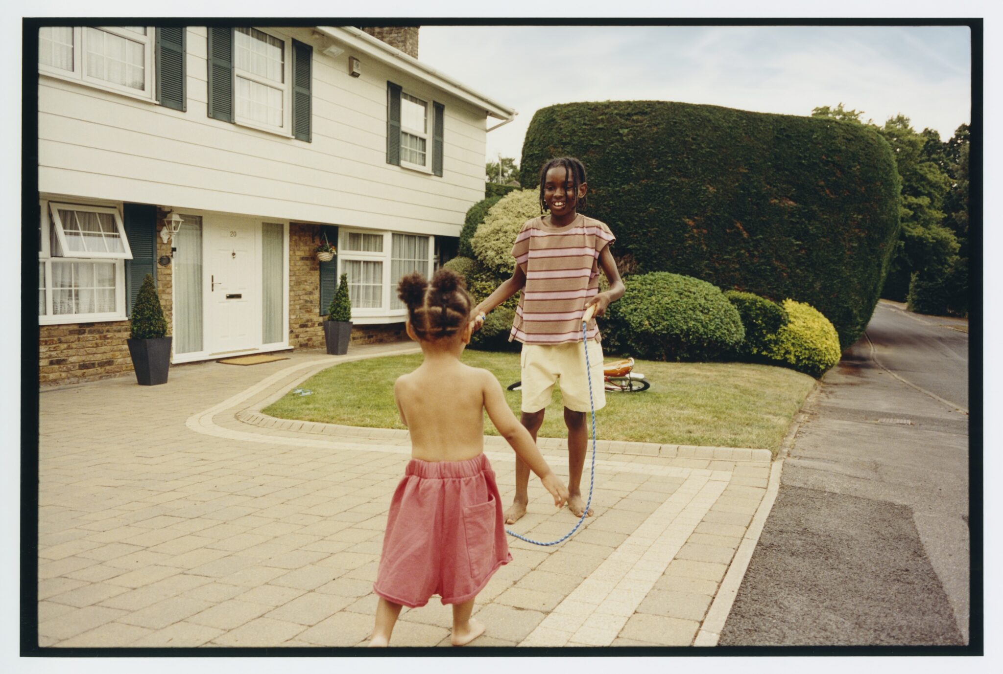 Child playing jump rope in a suburban driveway with younger toddler in pink shorts, striped summer outfit from MAIN STORY SS26 kids fashion editorial featured in Scimparello Magazine.