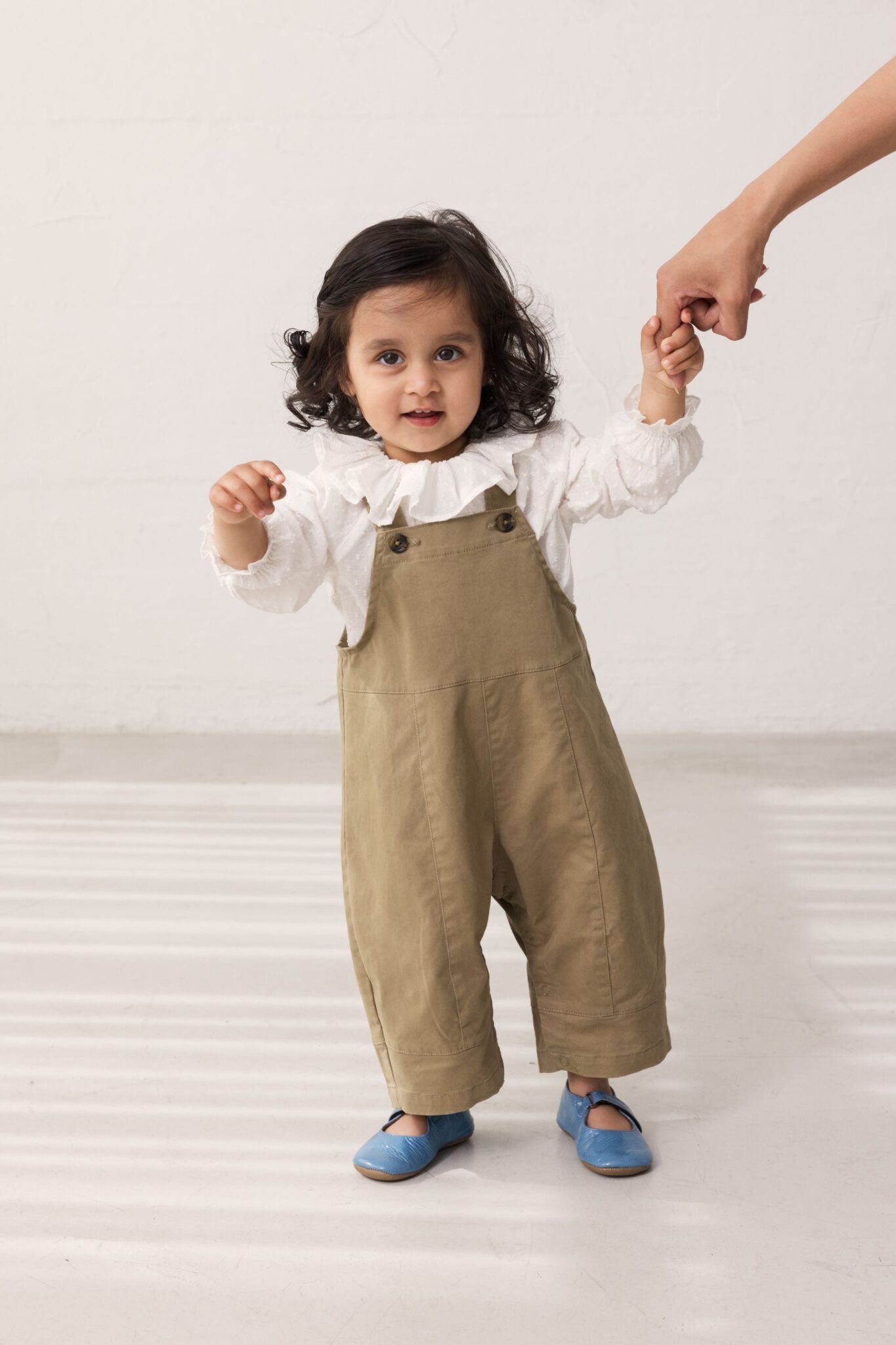 MarMar Copenhagen SS26 toddler collection featured in Scimparello Magazine – little girl learning to walk while holding an adult’s hand, wearing khaki cotton overalls over a white ruffled blouse and blue Mary Jane shoes in a neutral studio setting.