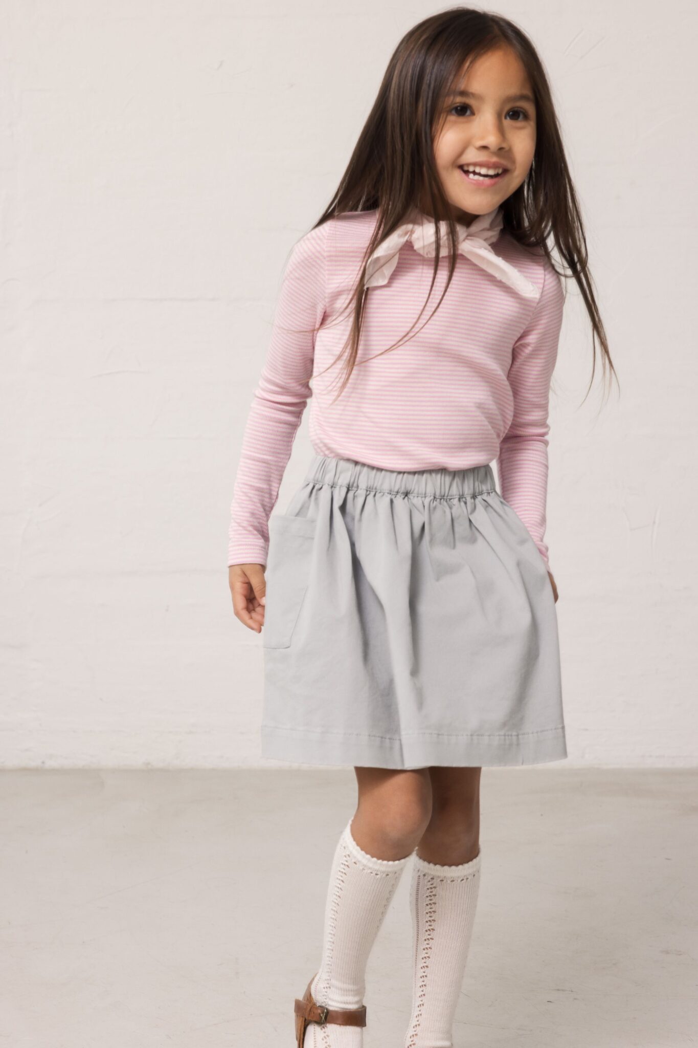 MarMar Copenhagen SS26 girls collection featured in Scimparello Magazine – smiling girl wearing a pink striped long-sleeve top with bow detail, light grey gathered skirt, knee-high socks and brown shoes against a minimal wall.