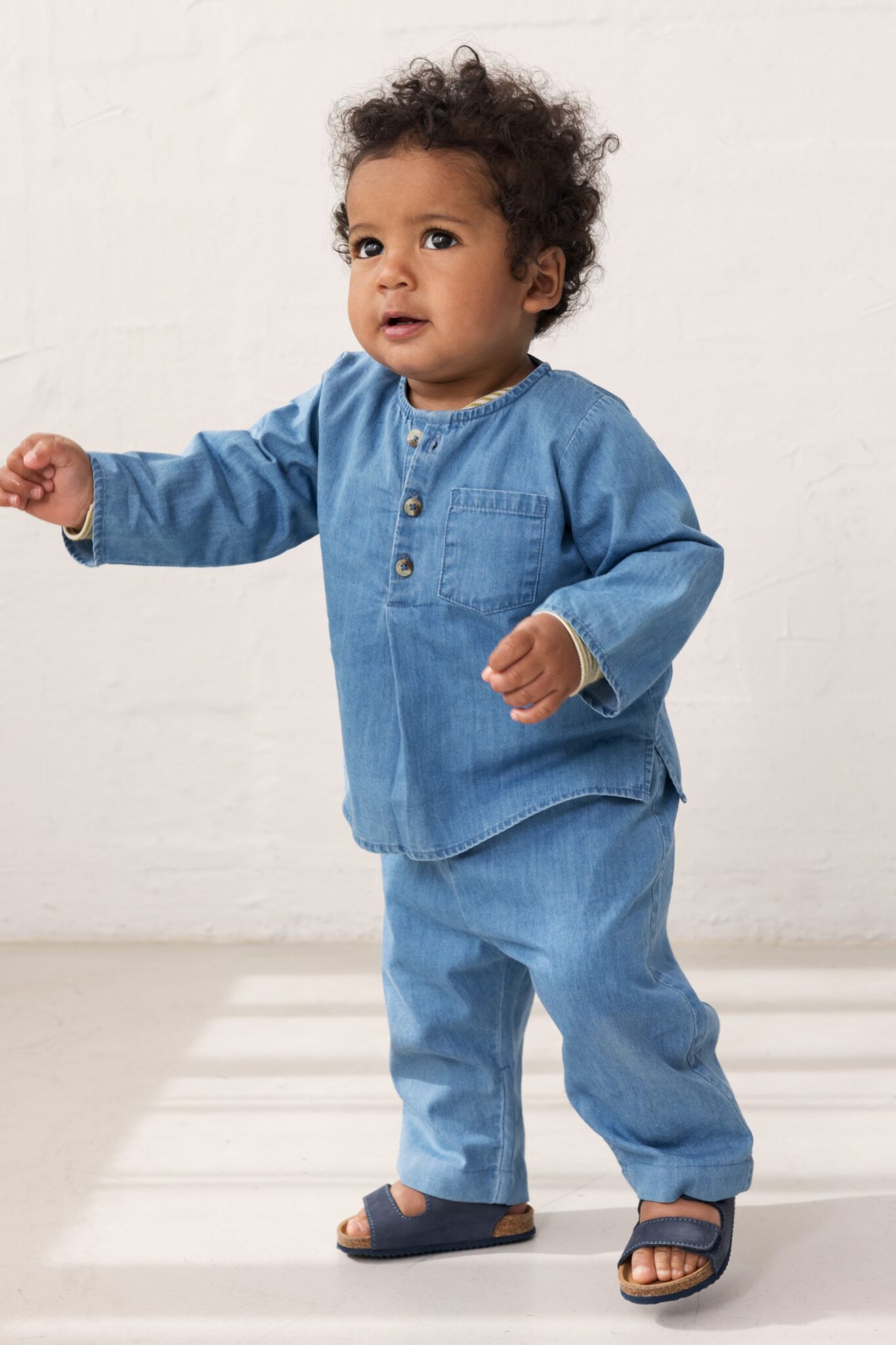 MarMar Copenhagen SS26 boys collection featured in Scimparello Magazine – toddler walking in a light denim two-piece set with long-sleeve top and matching trousers, styled with blue sandals in a minimal studio space.