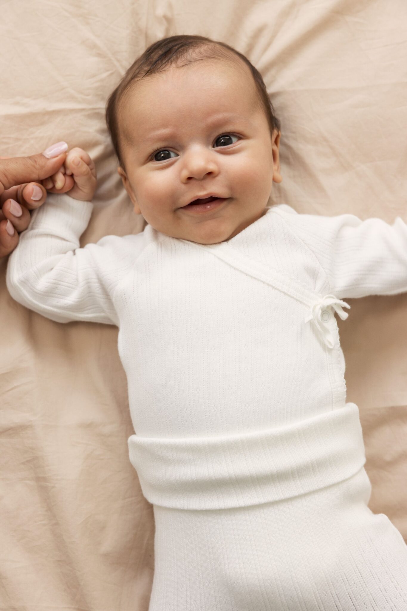 MarMar Copenhagen SS26 baby essentials featured in Scimparello Magazine – baby lying on a beige blanket wearing a soft white wrap-style bodysuit, holding an adult’s hand in warm natural light.