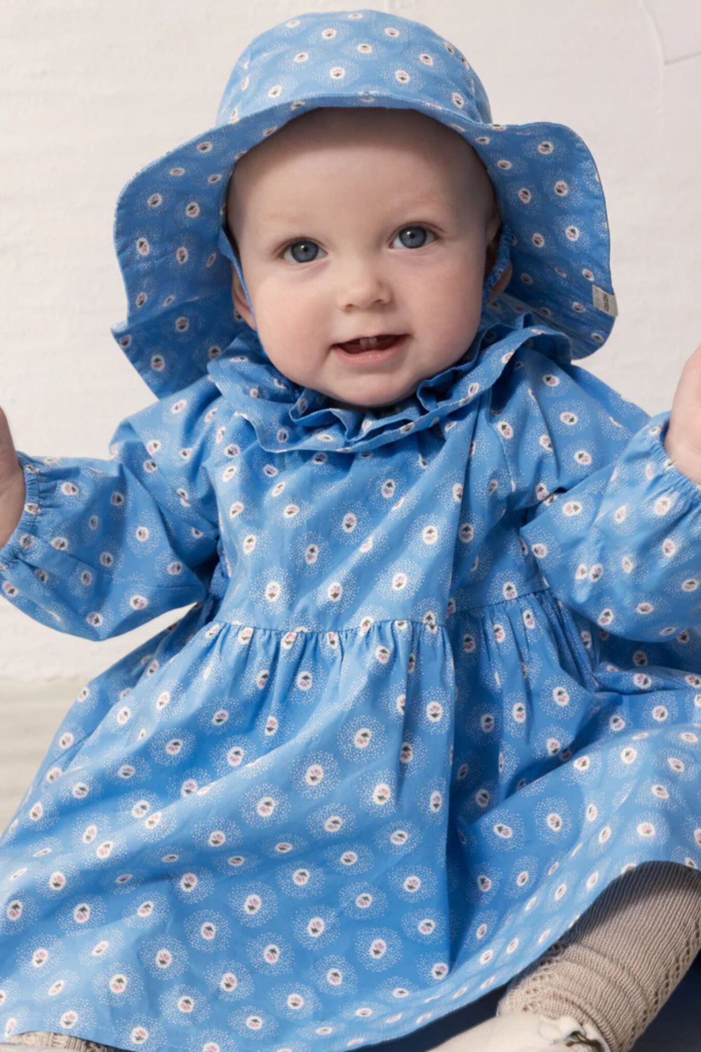 MarMar Copenhagen SS26 baby collection featured in Scimparello Magazine – baby sitting and smiling in a blue floral dress with matching wide-brim hat, delicate small flower print and soft neutral background.