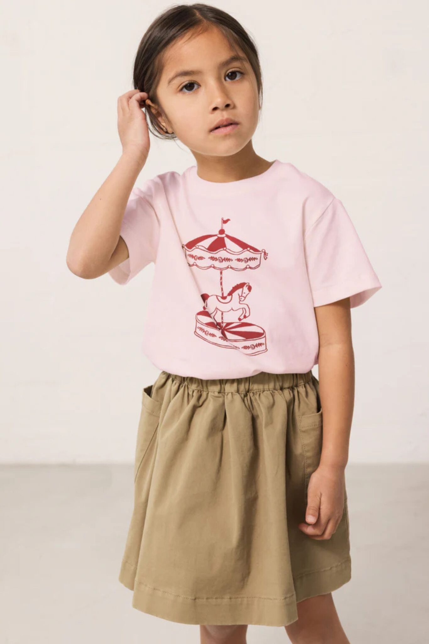 MarMar Copenhagen SS26 girls collection featured in Scimparello Magazine – girl wearing a light pink T-shirt with red carousel print paired with a khaki skirt, styled in a clean minimal studio setting.