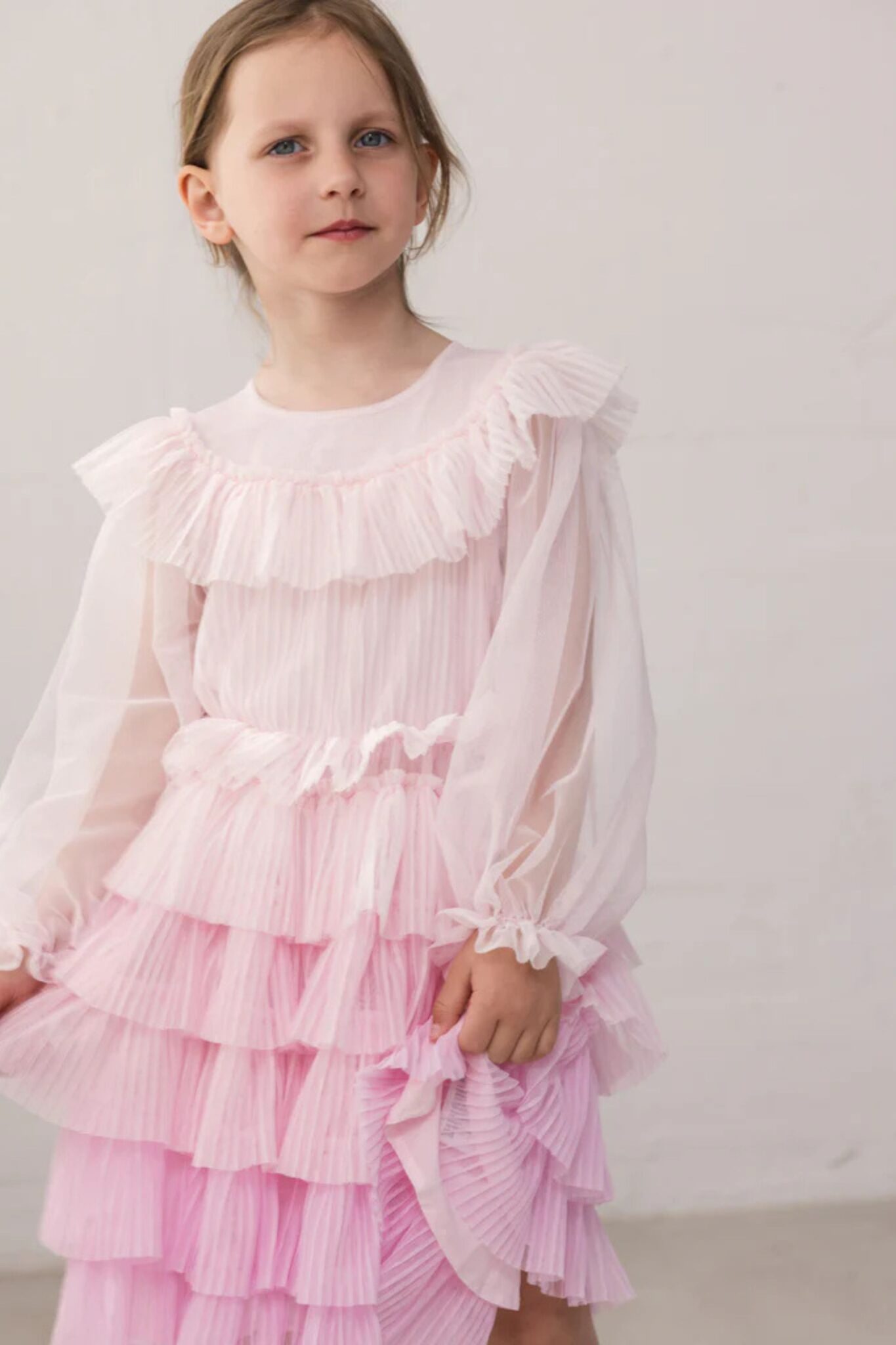 MarMar Copenhagen SS26 girls collection featured in Scimparello Magazine – girl wearing a soft pink pleated tulle dress with layered ruffles and sheer long sleeves, styled against a minimal neutral wall.