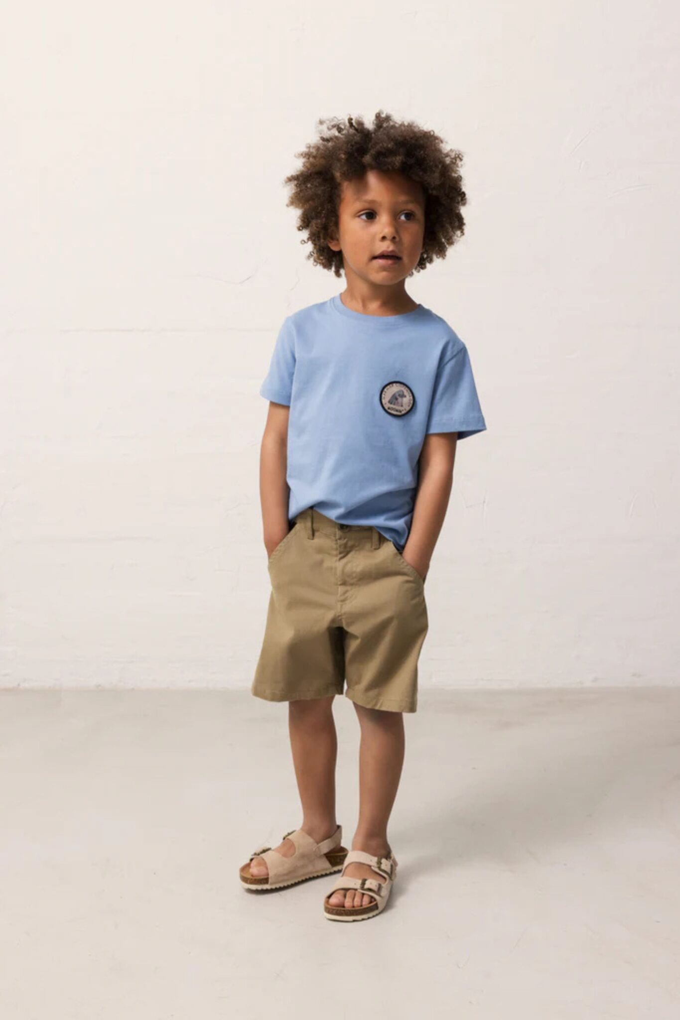 MarMar Copenhagen SS26 boys collection featured in Scimparello Magazine – young boy in a light blue T-shirt with small chest patch, khaki shorts and beige sandals, standing in a clean studio setting.