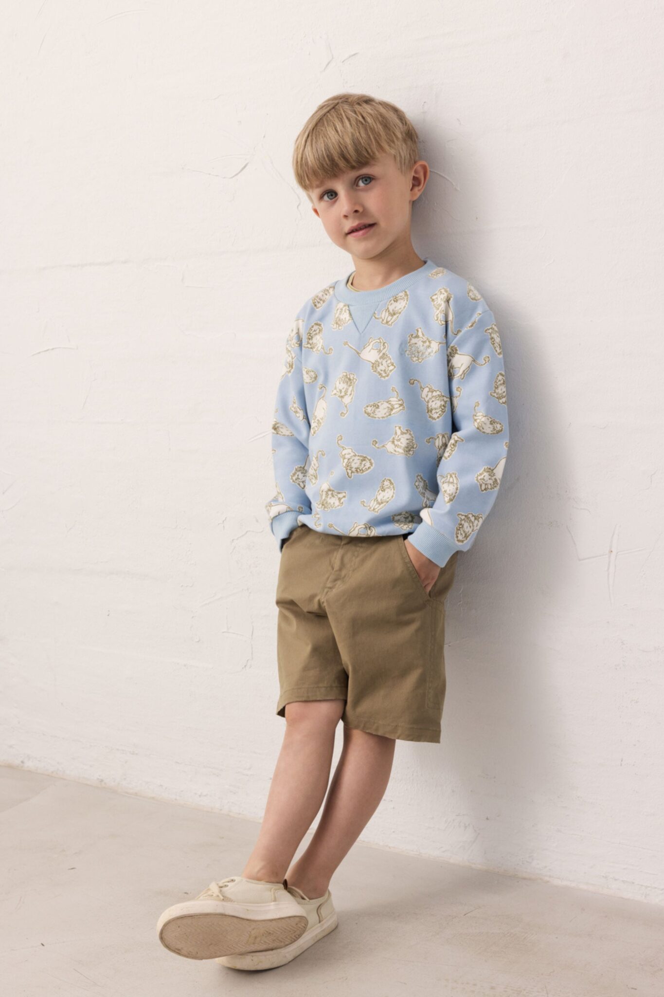 MarMar Copenhagen SS26 boys collection featured in Scimparello Magazine – boy wearing a light blue sweatshirt with playful print and khaki shorts, leaning against a textured white wall.