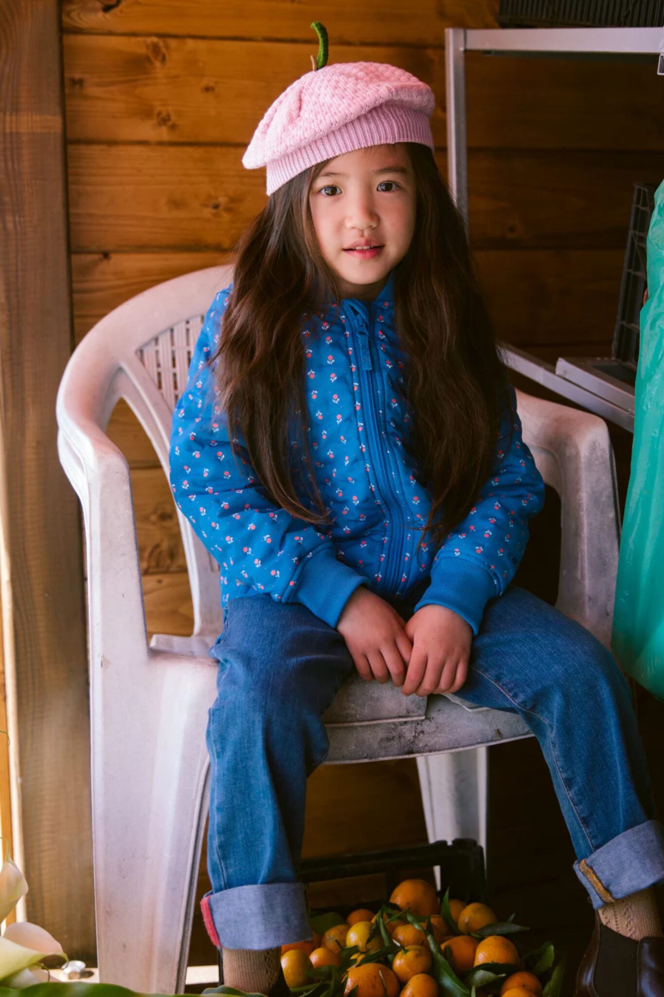 MarMar Copenhagen SS26 kids collection featured in Scimparello Magazine – girl sitting on a white chair in a wooden setting, wearing a blue floral zip-up jacket, rolled-up jeans and a soft pink knitted beret.