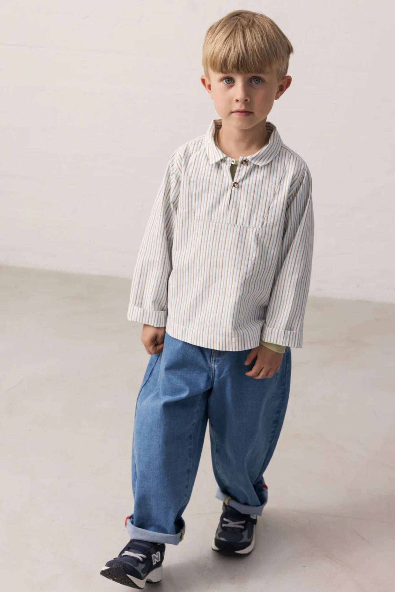 MarMar Copenhagen SS26 boys collection featured in Scimparello Magazine – young boy walking in a striped long-sleeve shirt with collar, relaxed blue jeans and navy sneakers in a minimal studio space.