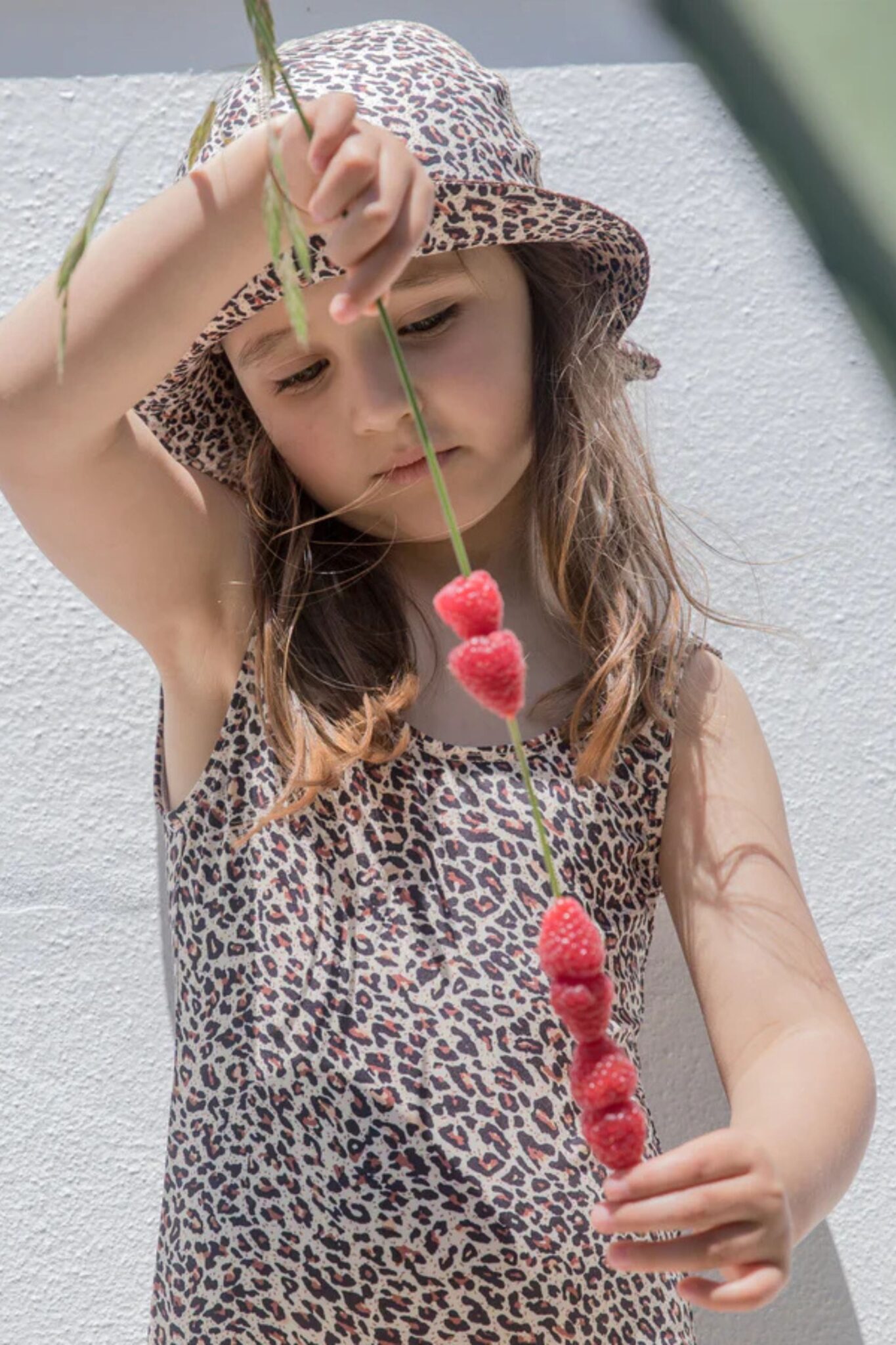MarMar Copenhagen SS26 girls collection featured in Scimparello Magazine – girl in a sleeveless leopard print dress and matching bucket hat holding a string of fresh raspberries against a sunlit white wall.