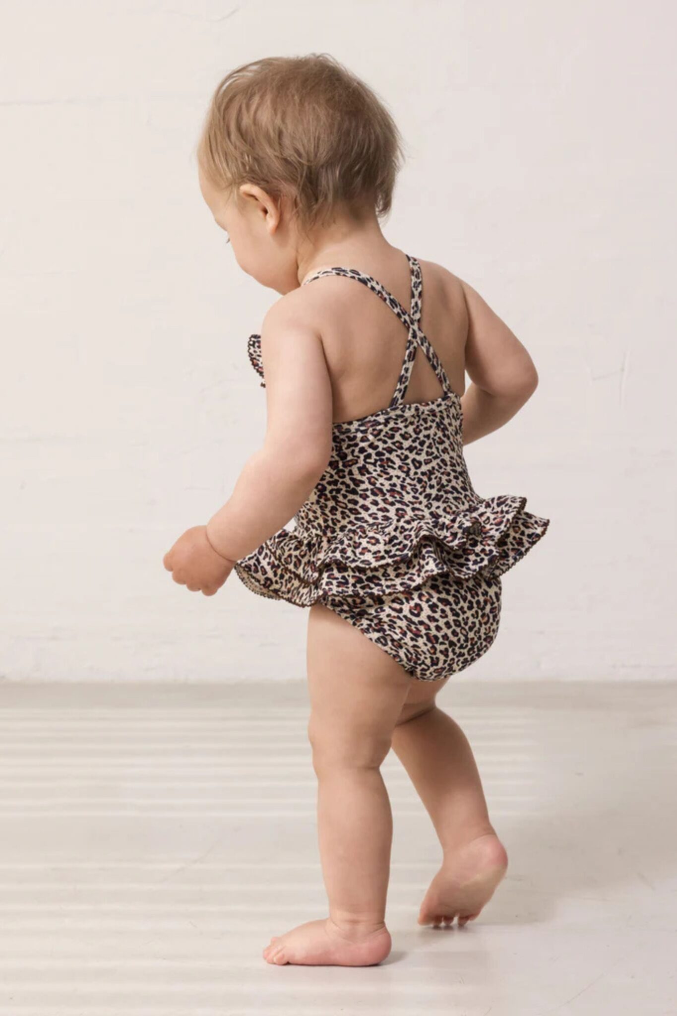 MarMar Copenhagen SS26 baby collection featured in Scimparello Magazine – toddler walking away wearing a leopard print romper with crossed back straps and ruffle detail.
