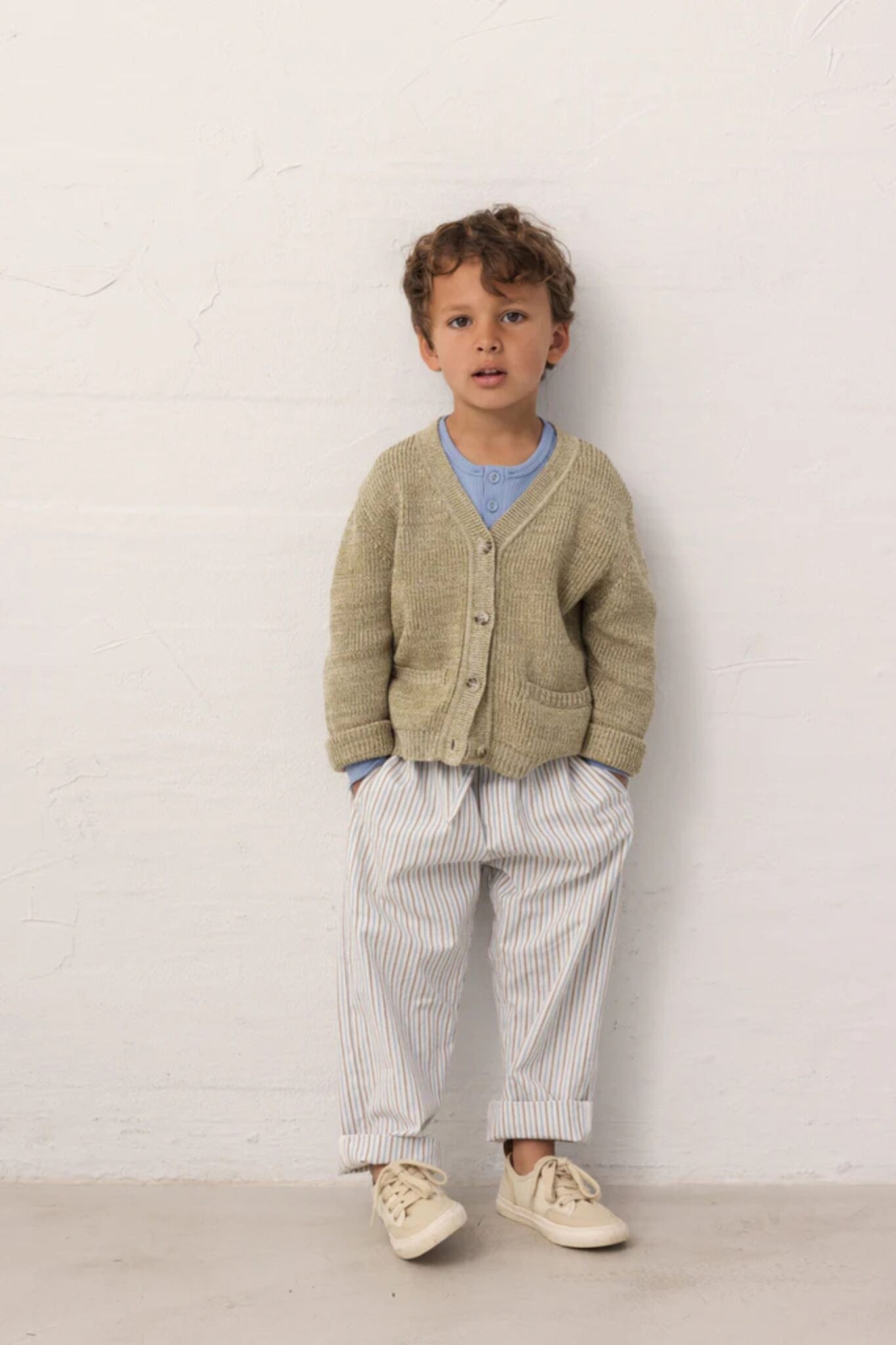 MarMar Copenhagen SS26 kids collection featured in Scimparello Magazine – young boy standing against a neutral textured wall wearing a soft beige knitted cardigan layered over a light blue henley shirt, striped cotton trousers with rolled cuffs, and cream lace-up sneakers in natural tones.
