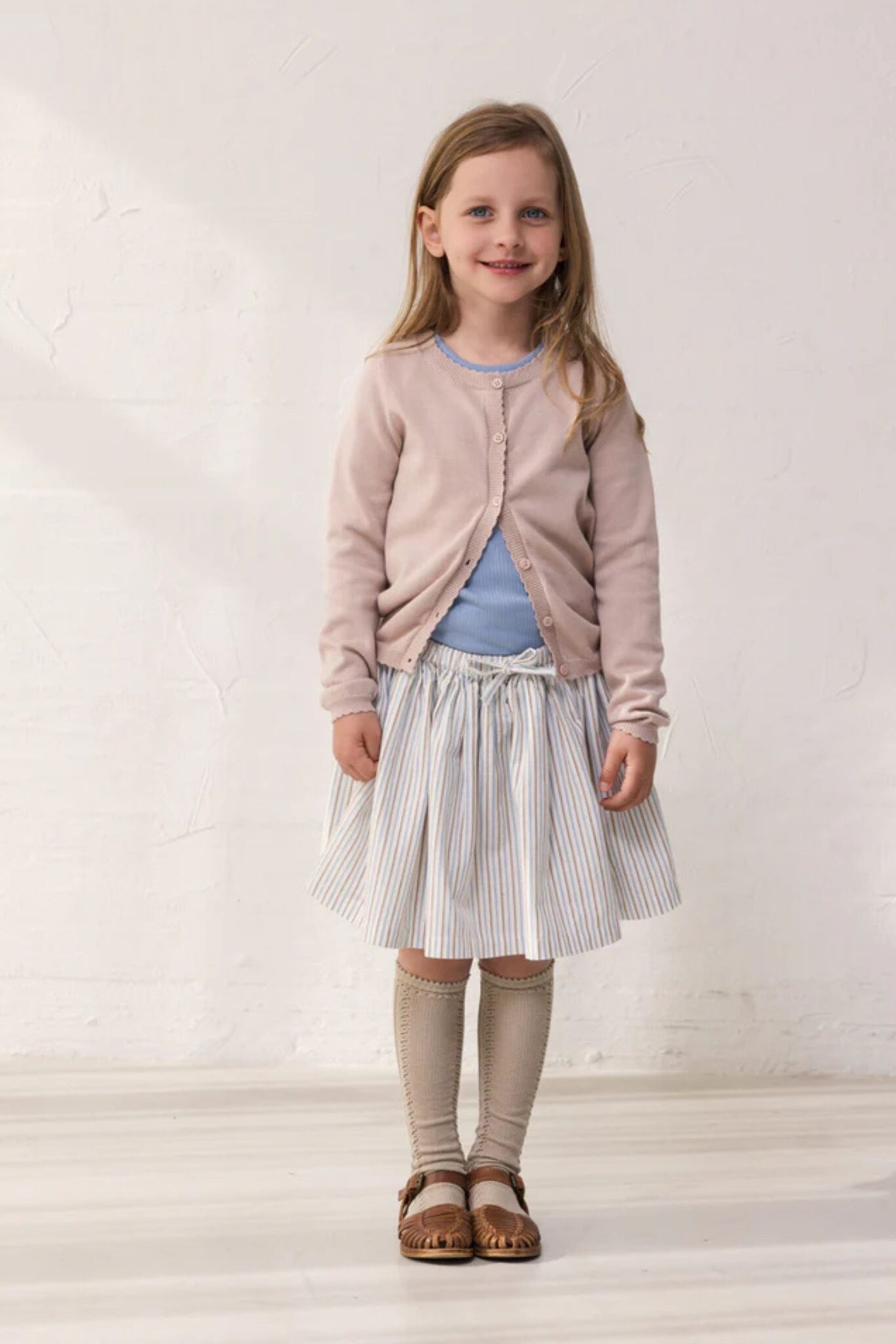 MarMar Copenhagen SS26 girls collection featured in Scimparello Magazine – girl in a soft pink cardigan over a light blue top paired with a striped skirt, knee-high socks and brown leather sandals.