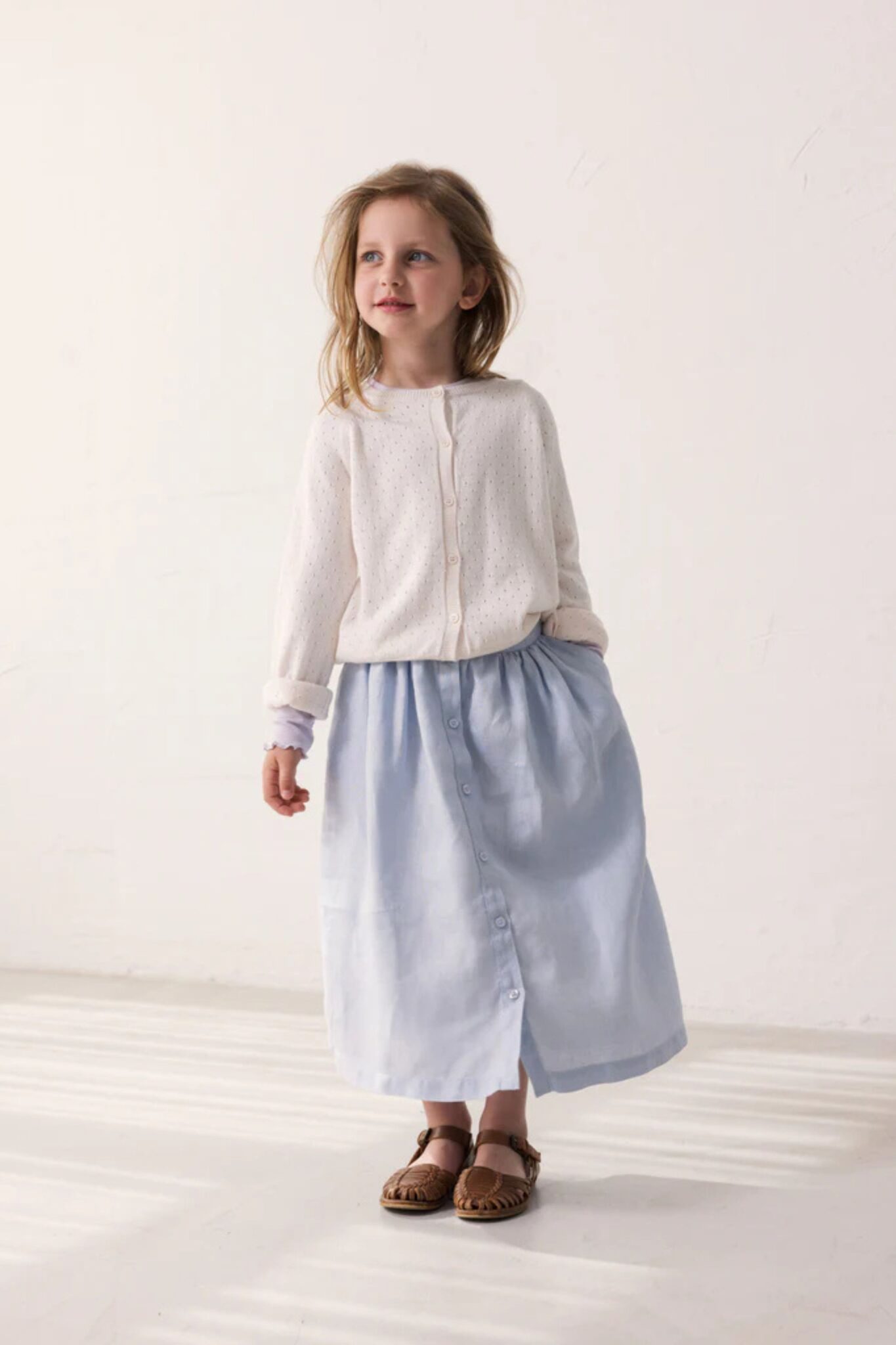 MarMar Copenhagen SS26 girls collection featured in Scimparello Magazine – girl wearing a white cardigan and light blue midi skirt with front buttons, styled with brown woven sandals.