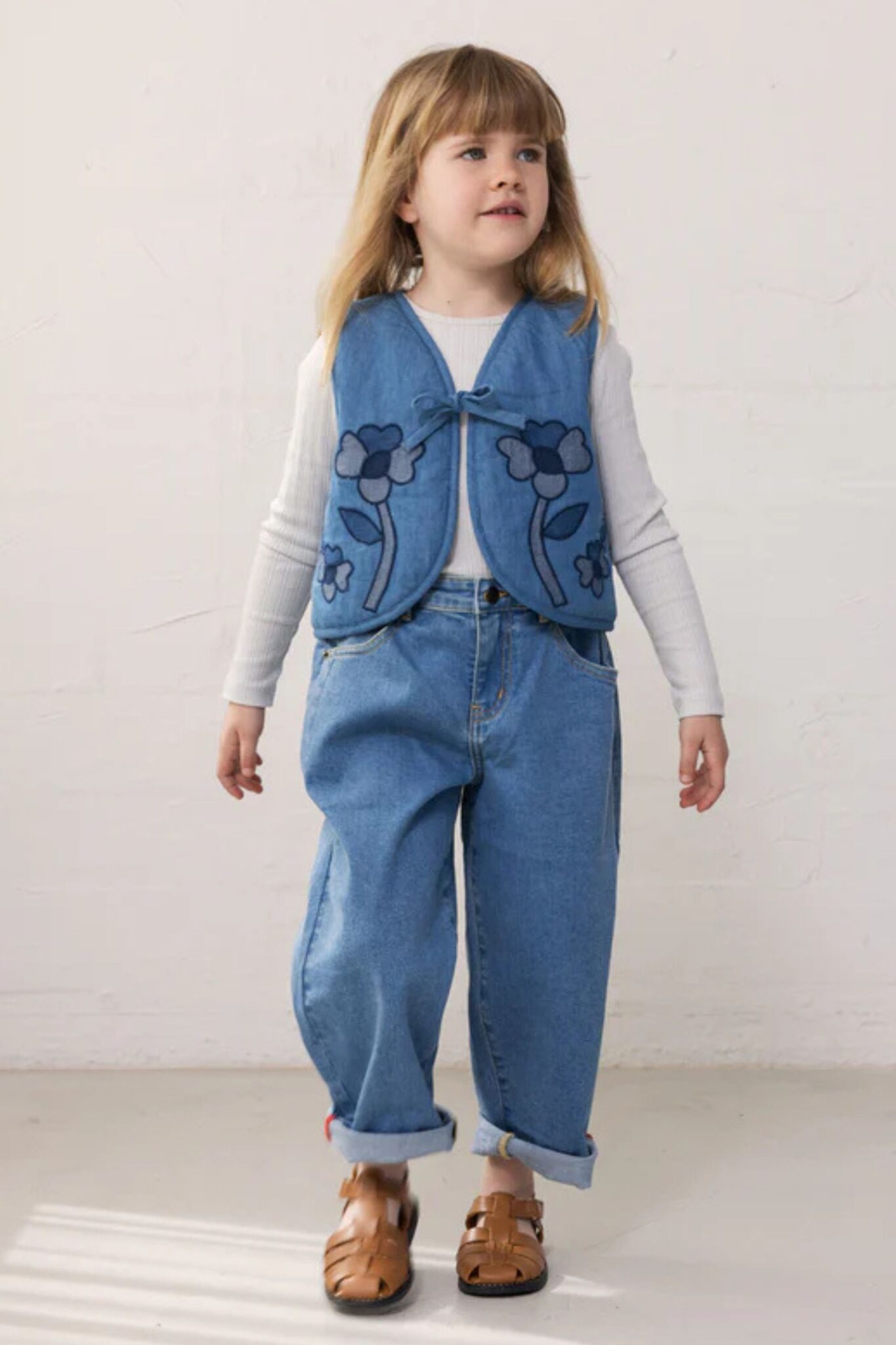 MarMar Copenhagen SS26 girls denim collection featured in Scimparello Magazine – girl in wide-leg jeans and a blue vest with floral appliqué over a white long-sleeve top, walking in a minimal studio setting.