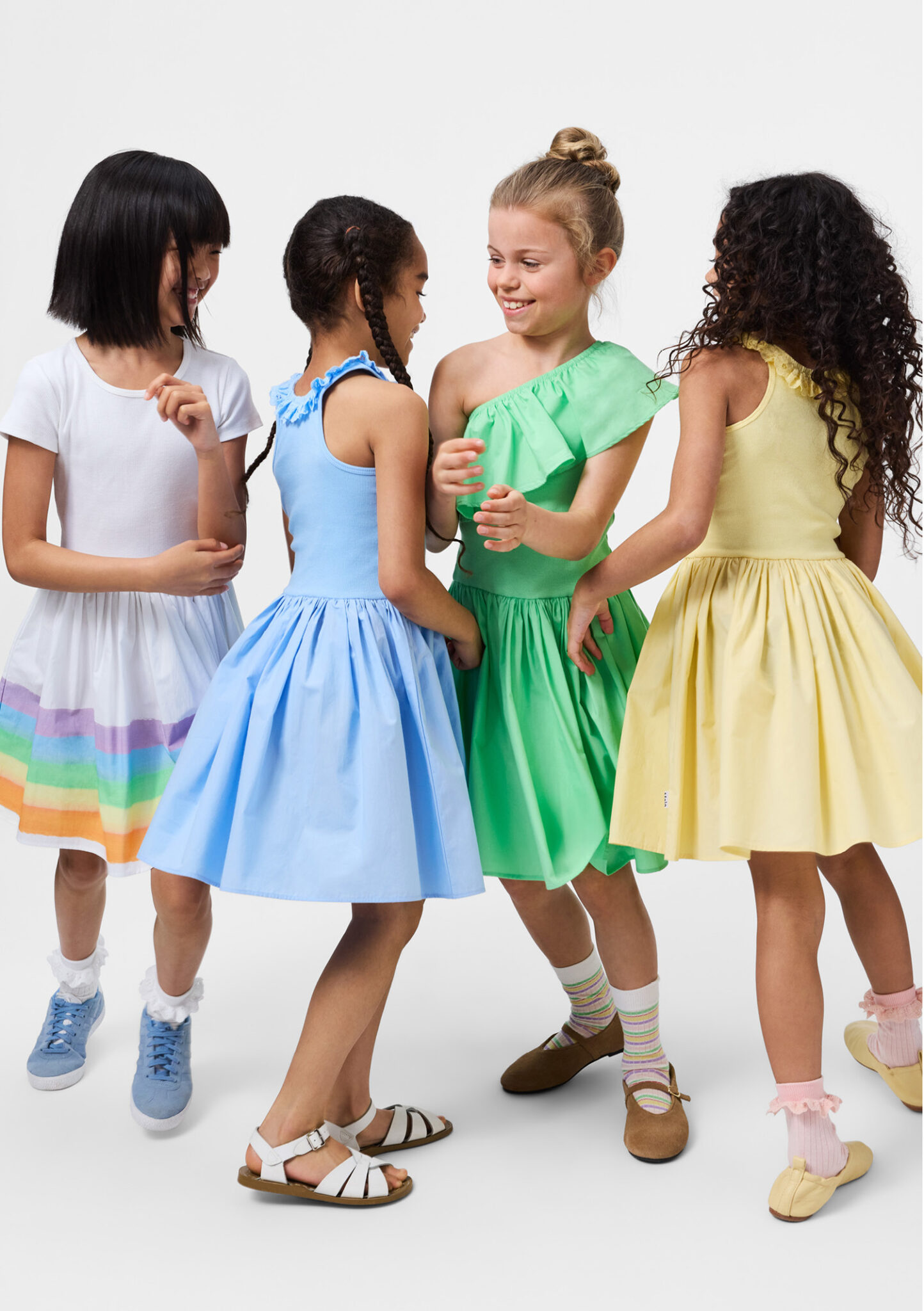 Four girls wearing colorful summer dresses in white, blue, green and yellow from Molo SS26, studio background, playful kids fashion featured in Scimparello Magazine.